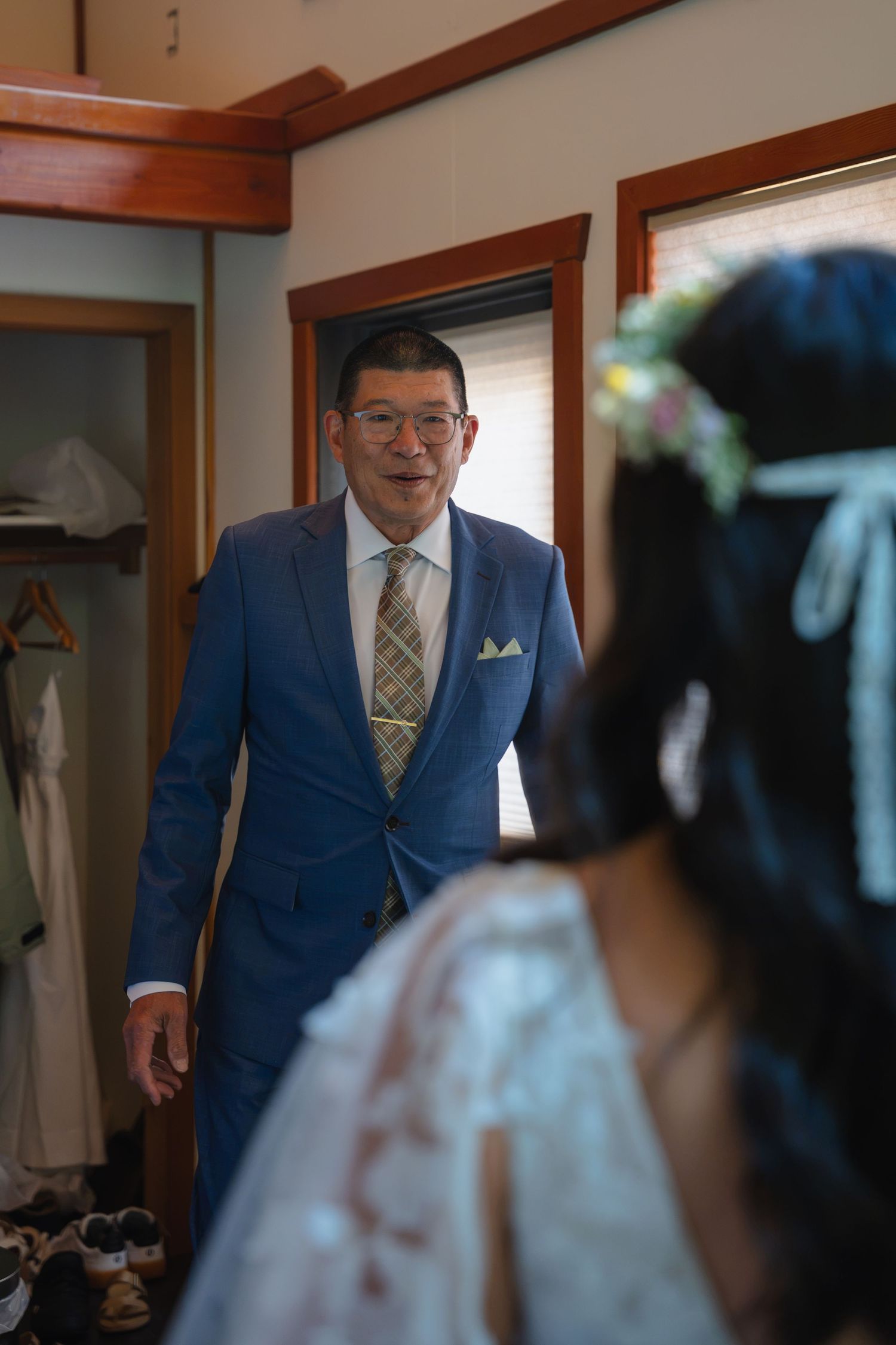 A man in a blue suit and tie smiles at a bride wearing a floral crown during wedding preparations.