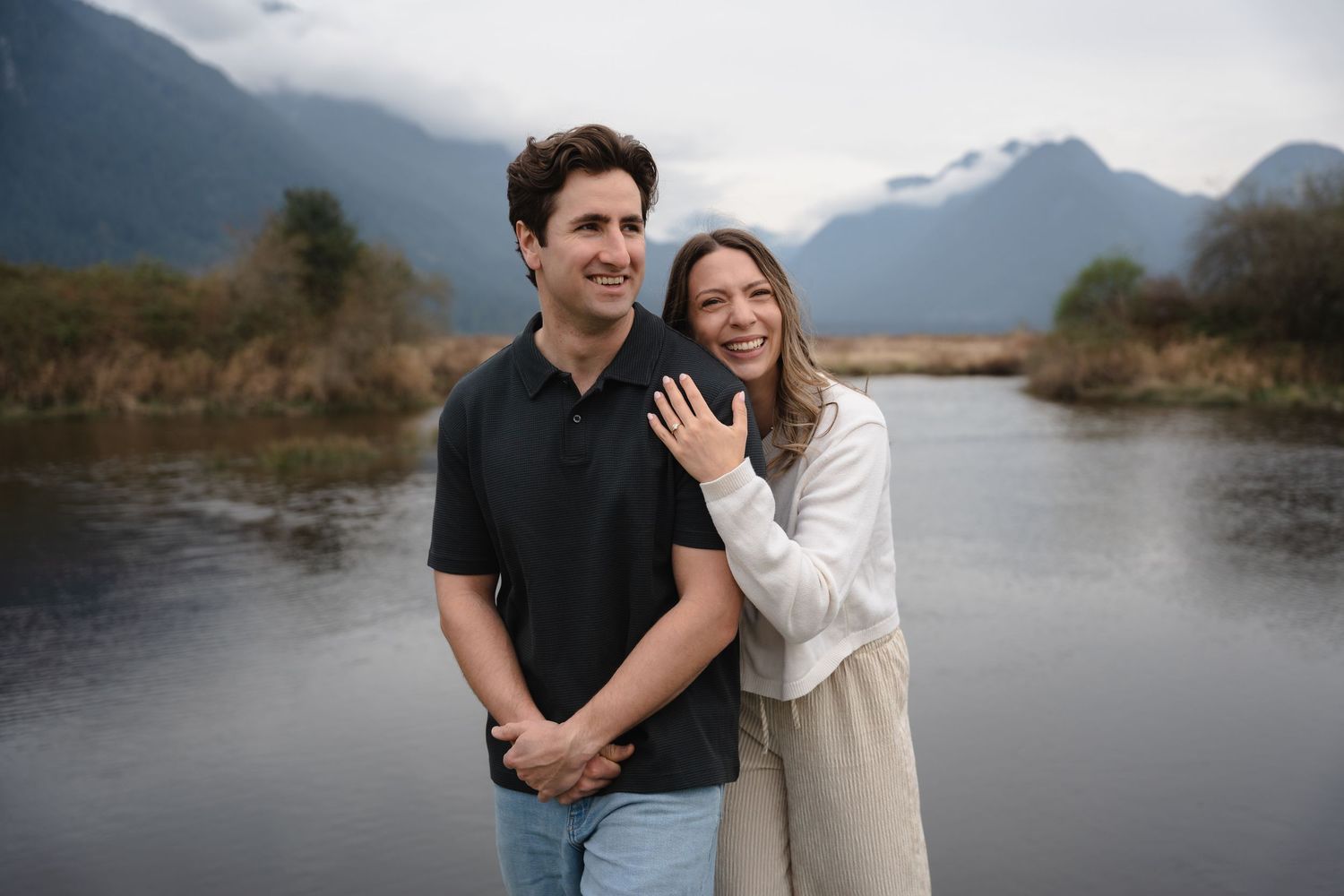 Happy couple embracing outdoors near a river with mountain backdrop, woman showing engagement ring.