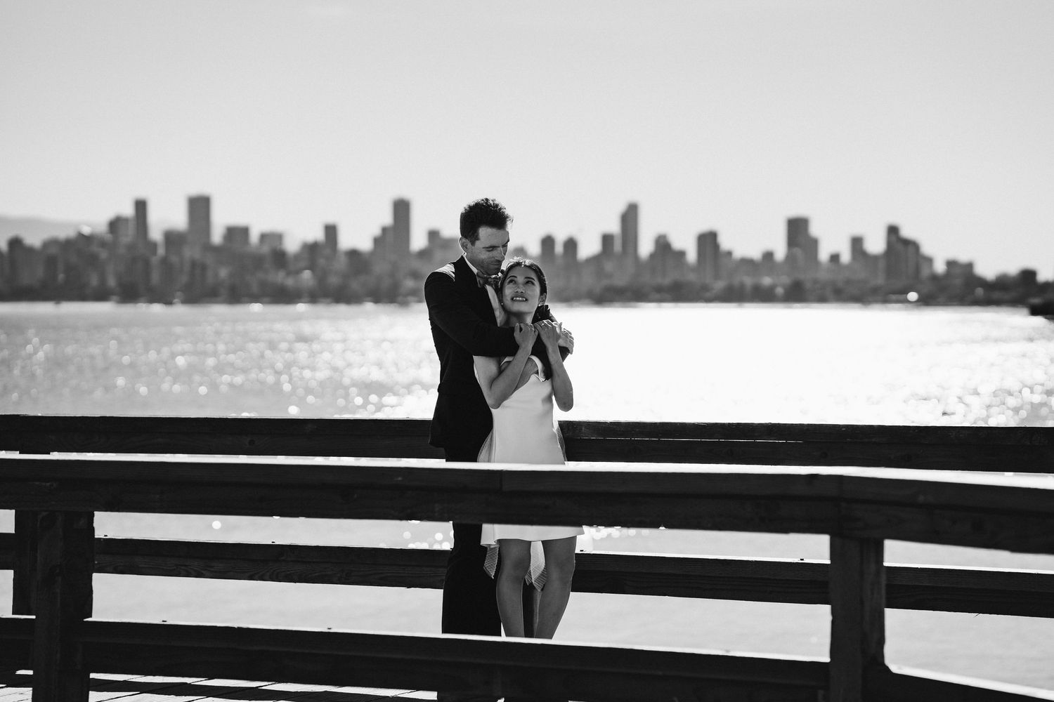 Couple embracing on a pier with a city skyline and sparkling water in the background, black and white photo.