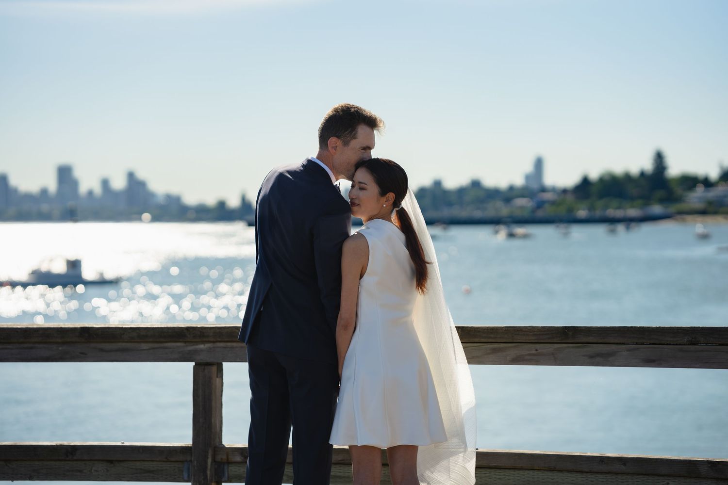 Bride and groom share a romantic moment on a waterfront pier with a city skyline backdrop on their wedding day.