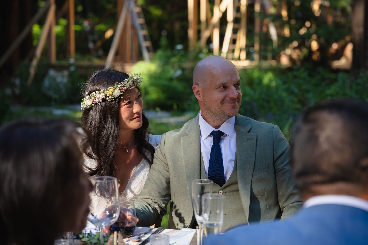 A bride wearing a floral crown and groom in sage suit smile at an outdoor wedding reception.