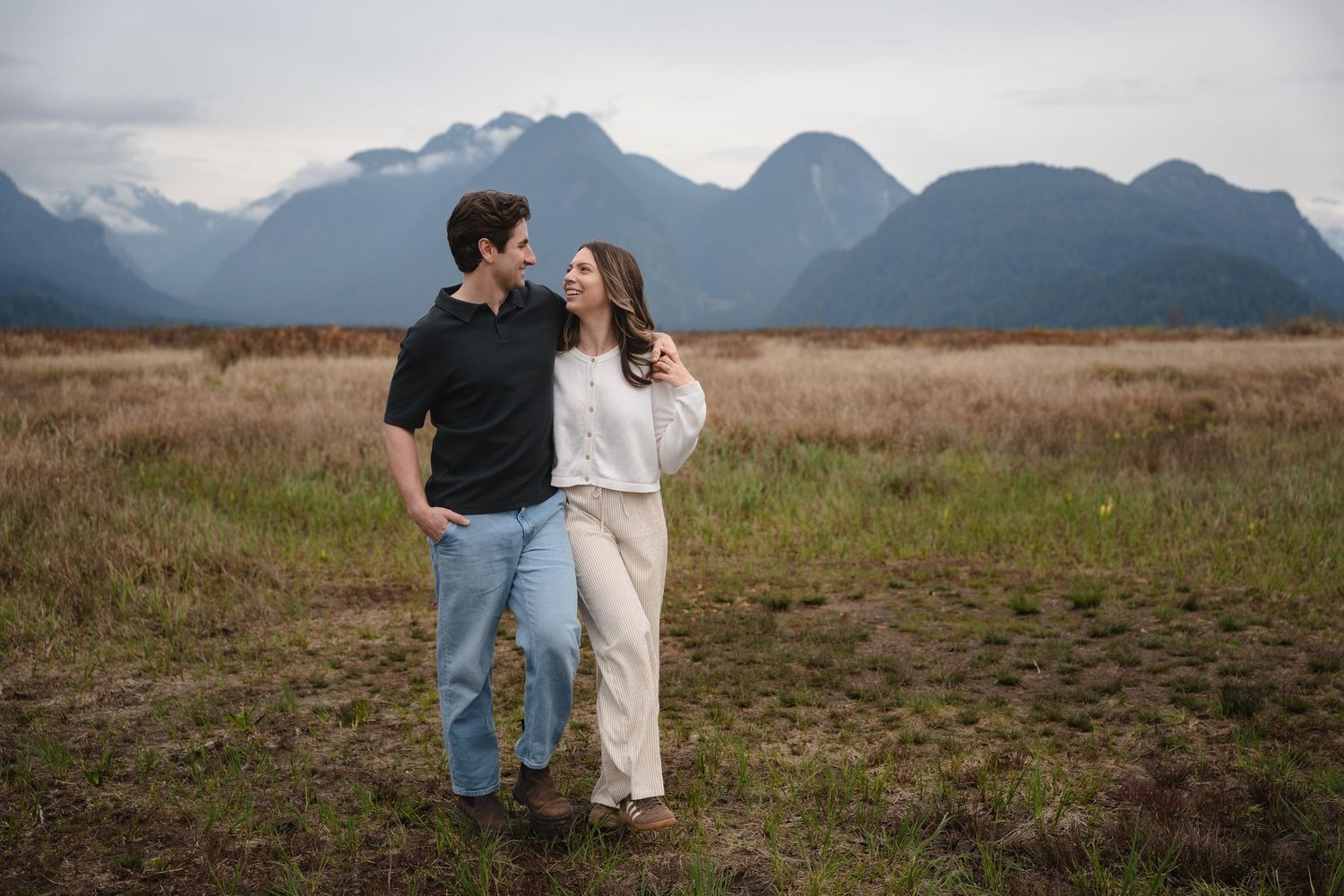 Couple embracing in open field with stunning mountain range backdrop on cloudy day.