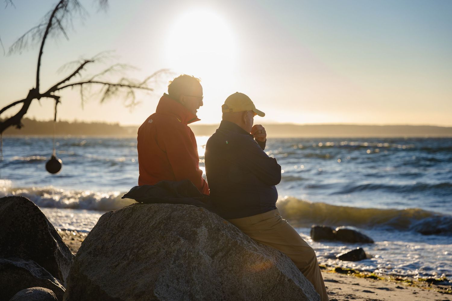 Two people sitting on rocks by the ocean shore at sunset, silhouetted against golden light.