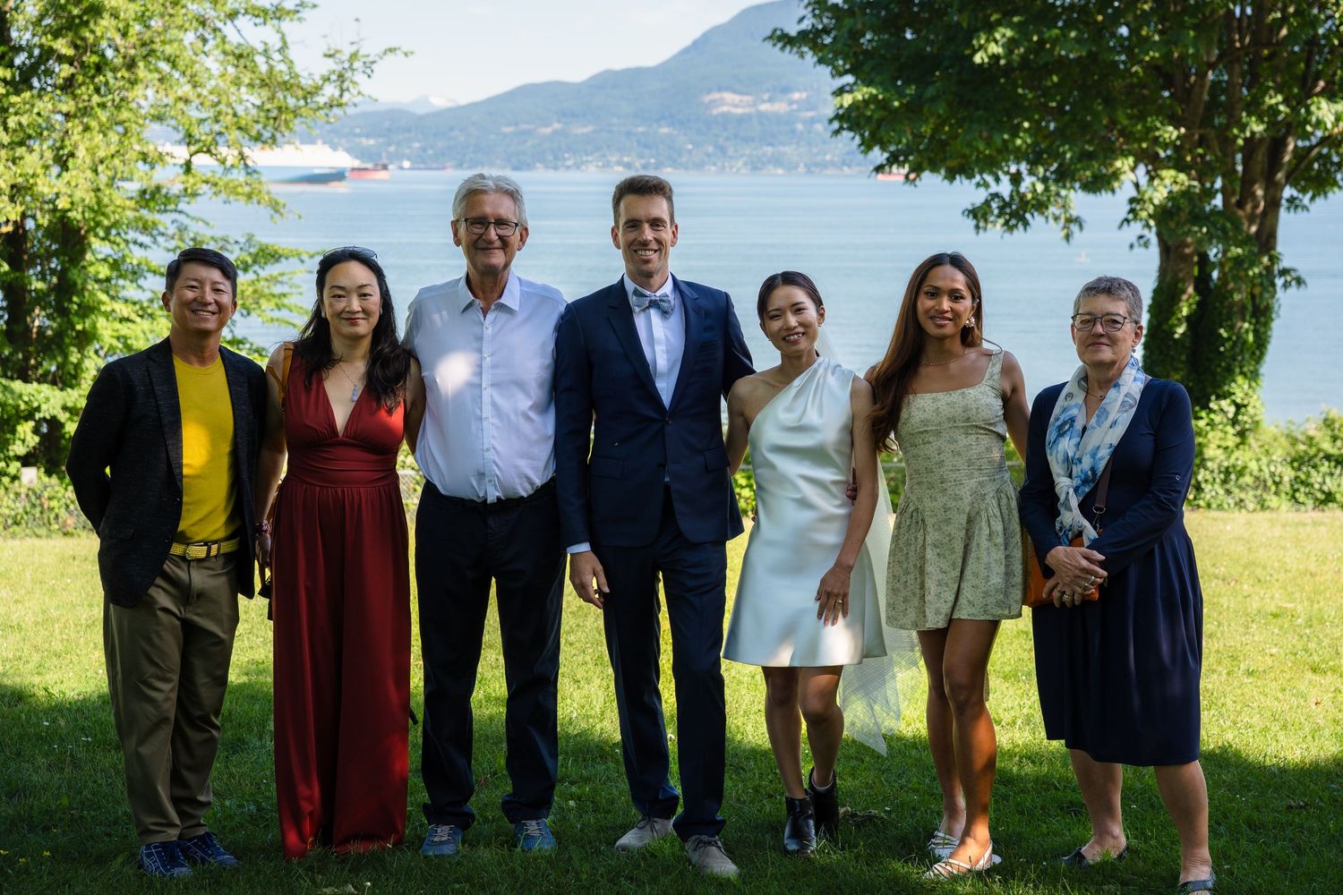 Group of seven people posing together outdoors with a scenic lake and mountains in the background.