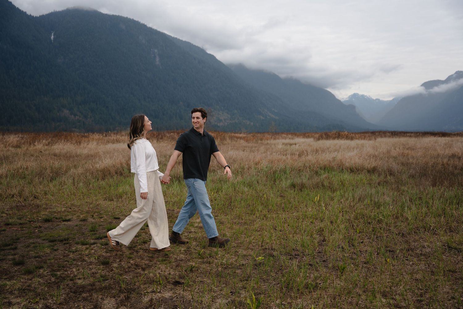 Couple holding hands walking through a scenic mountain meadow under cloudy skies in a romantic outdoor setting.