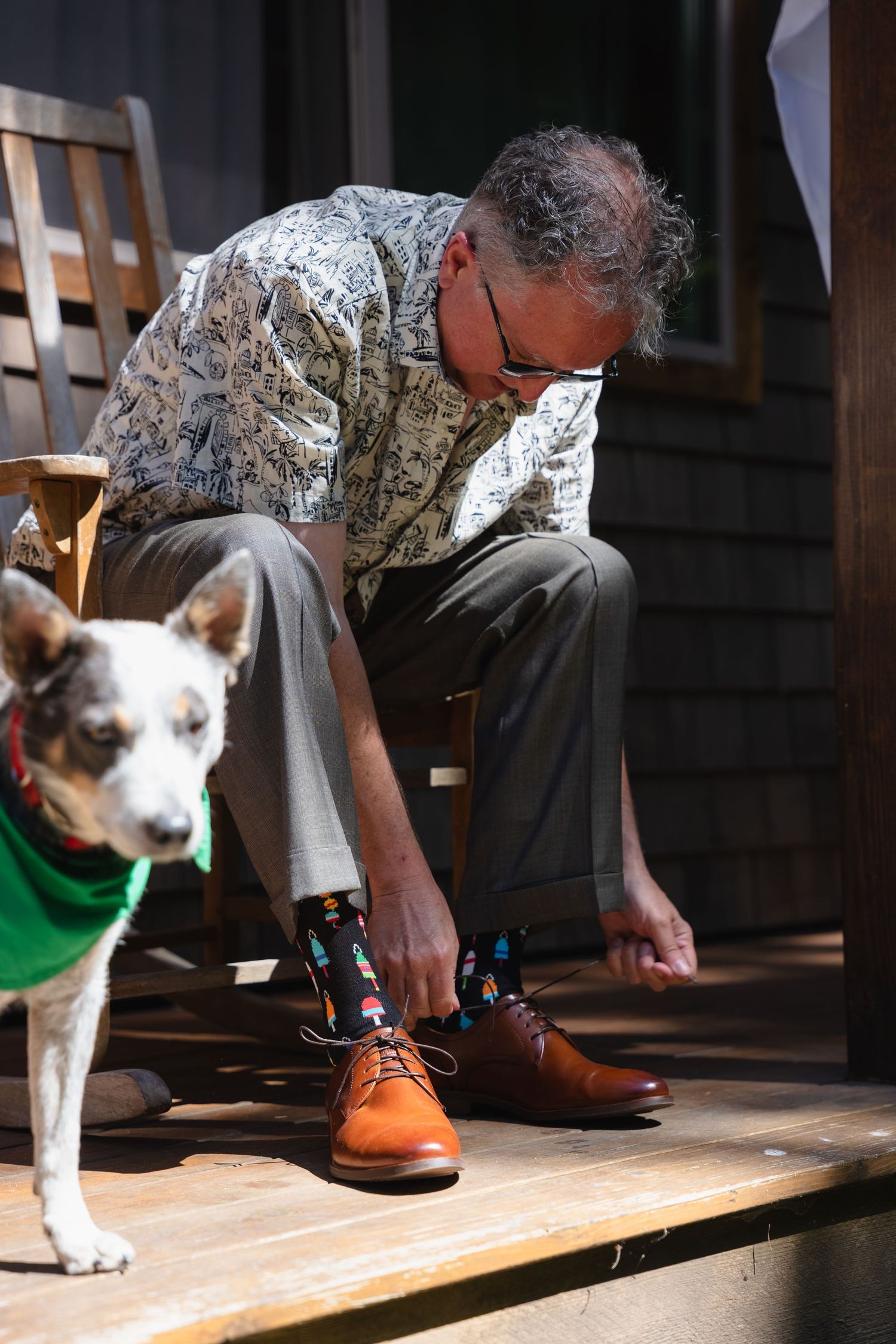 Man tying brown dress shoes with colorful socks on a porch beside a dog wearing a green bandana.