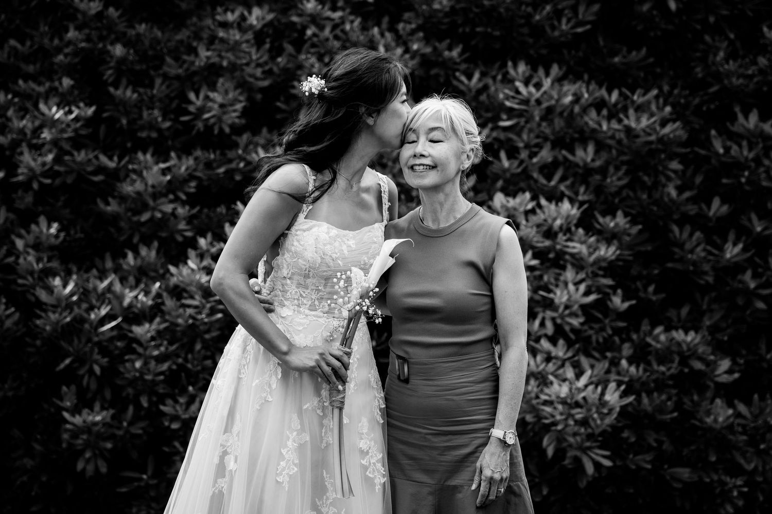 Black and white photo of bride in lace gown kissing an older woman on the cheek outdoors.