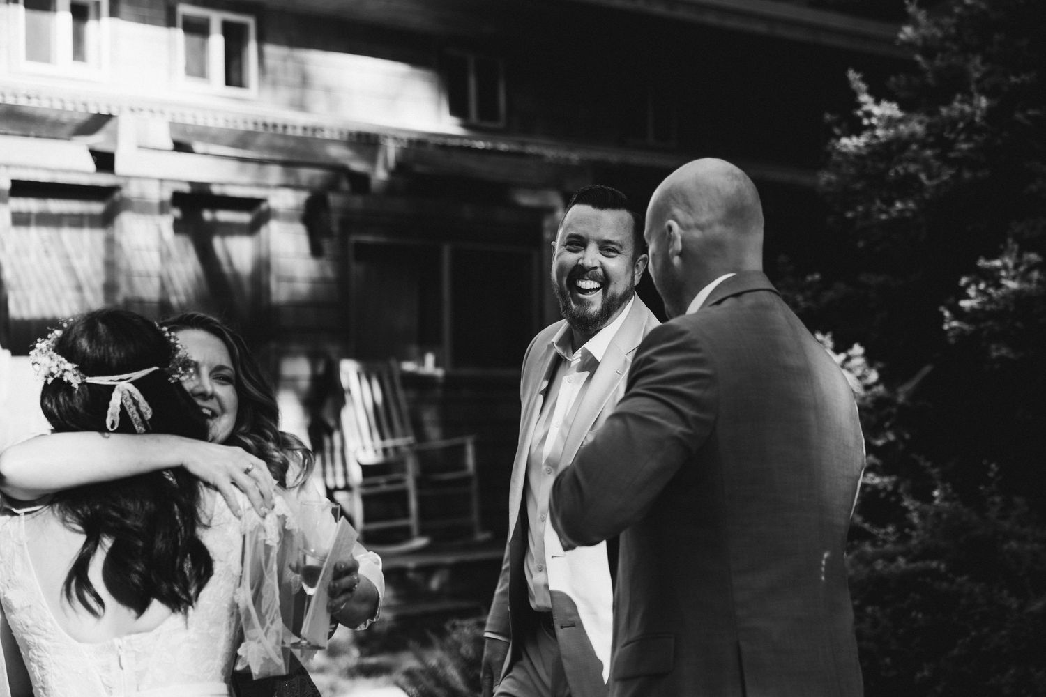 Black and white wedding photo of a groom laughing as guests embrace and celebrate outside a rustic venue.