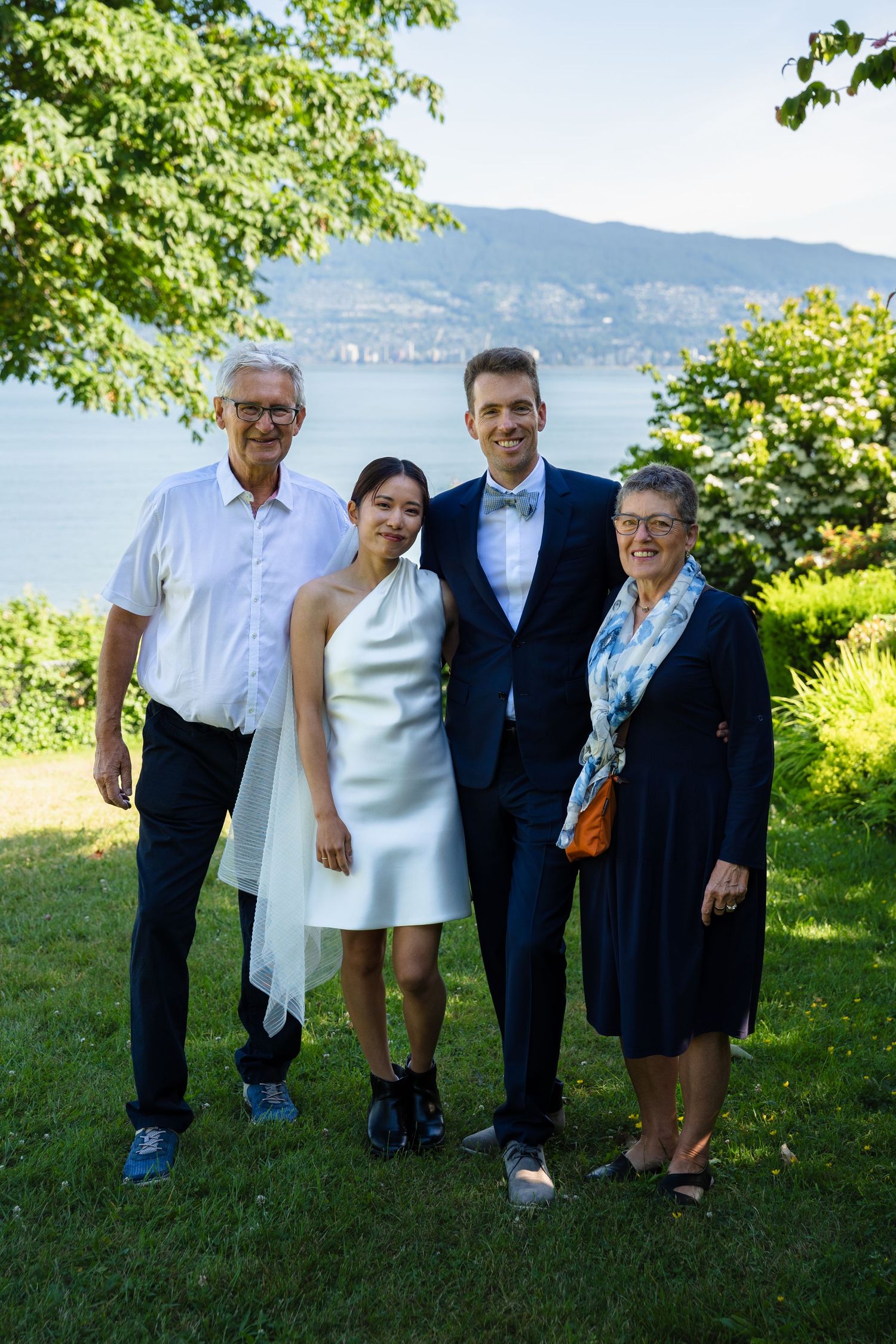 Wedding couple poses with family in a garden with scenic lake and mountain views on a sunny day.