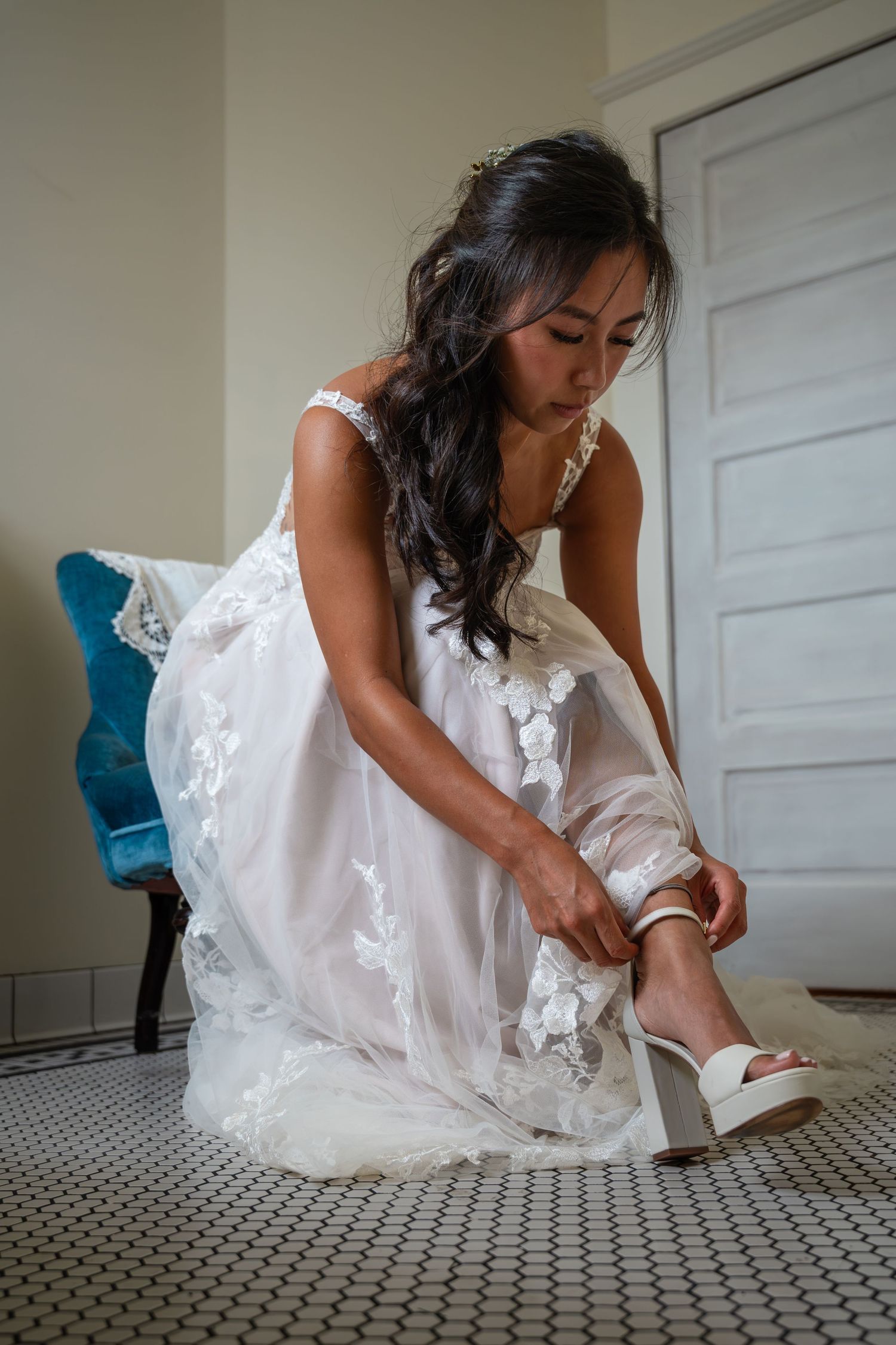Bride in white lace wedding gown fastening white heeled sandal while seated indoors.