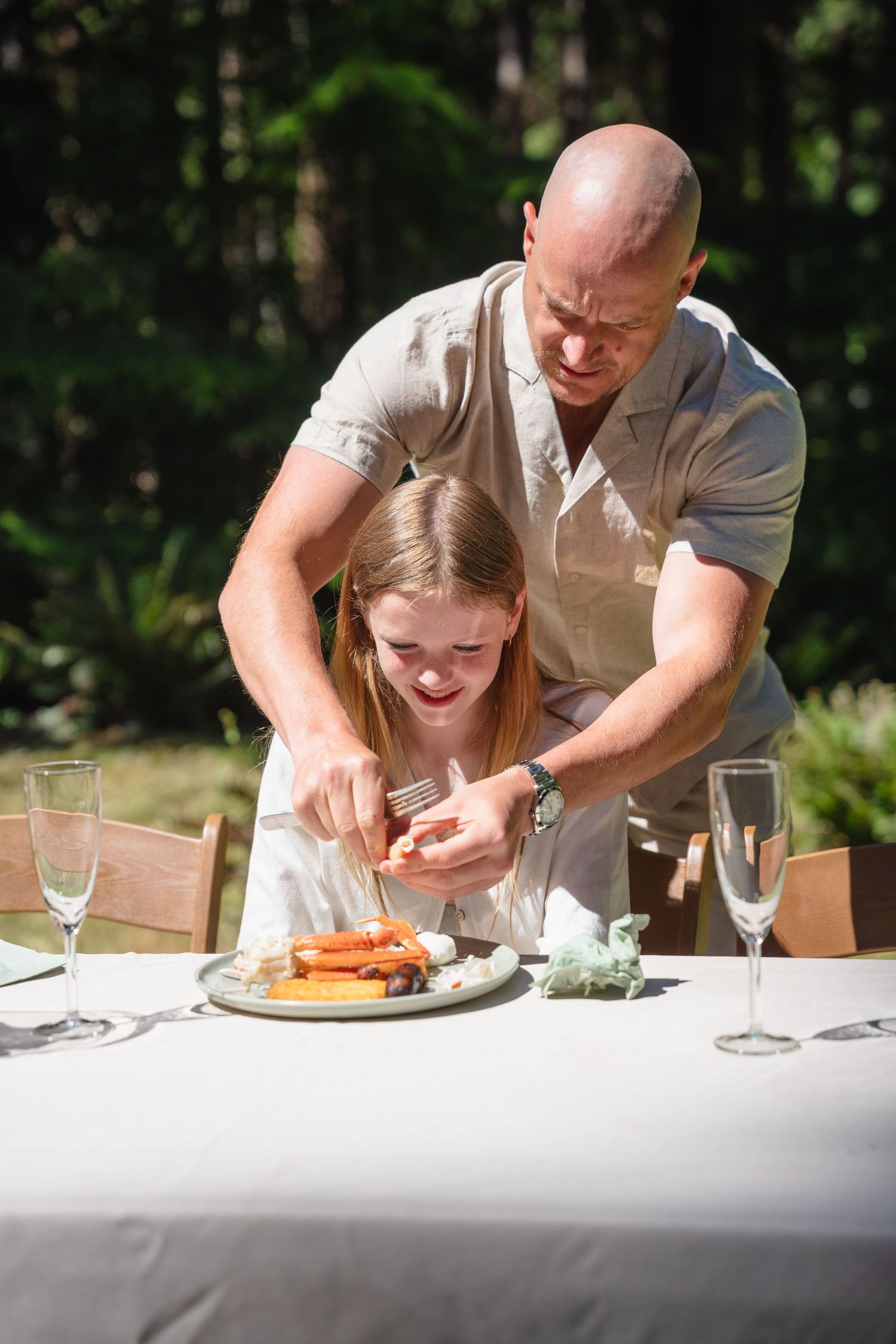 Bald father helps smiling daughter cut food at elegant outdoor dining table set with champagne flutes.