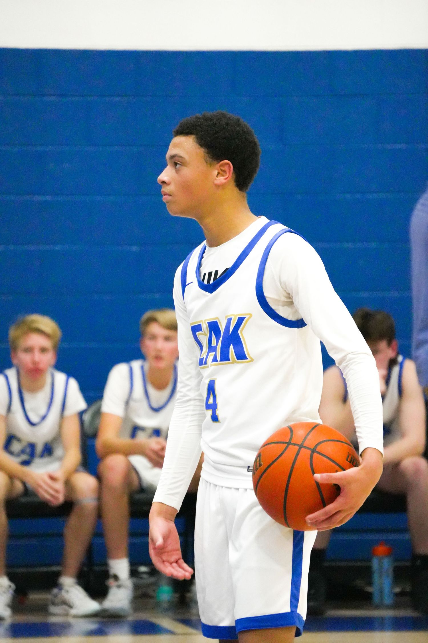 Basketball players in white jerseys stand on a blue court during practice with basketballs in hand.