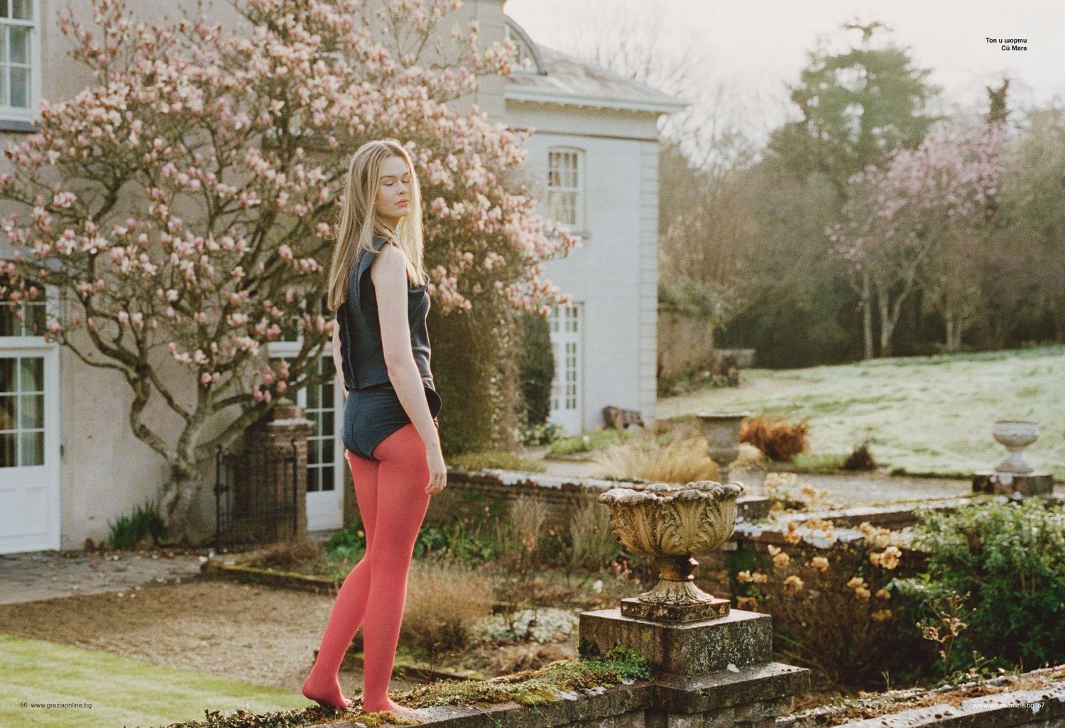 Young woman in red tights and navy top poses in a garden beside a blooming magnolia tree near a country house.