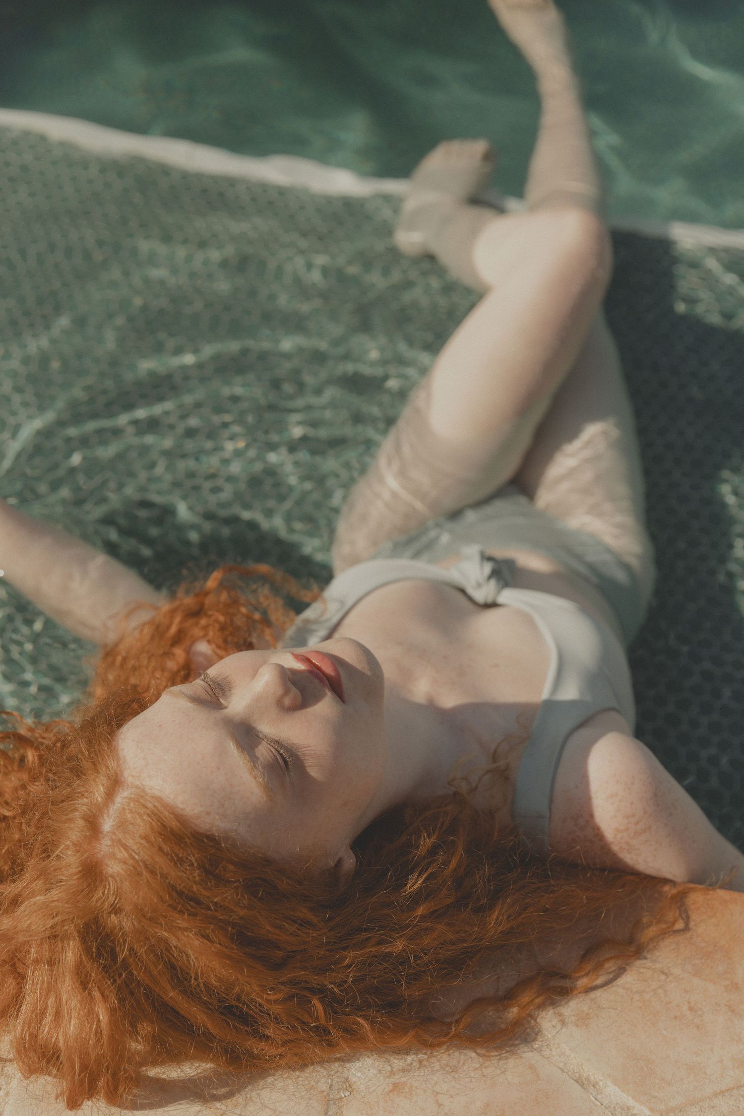 Editorial image of female model with red hair floats in pool wearing light-coloured swimwear.