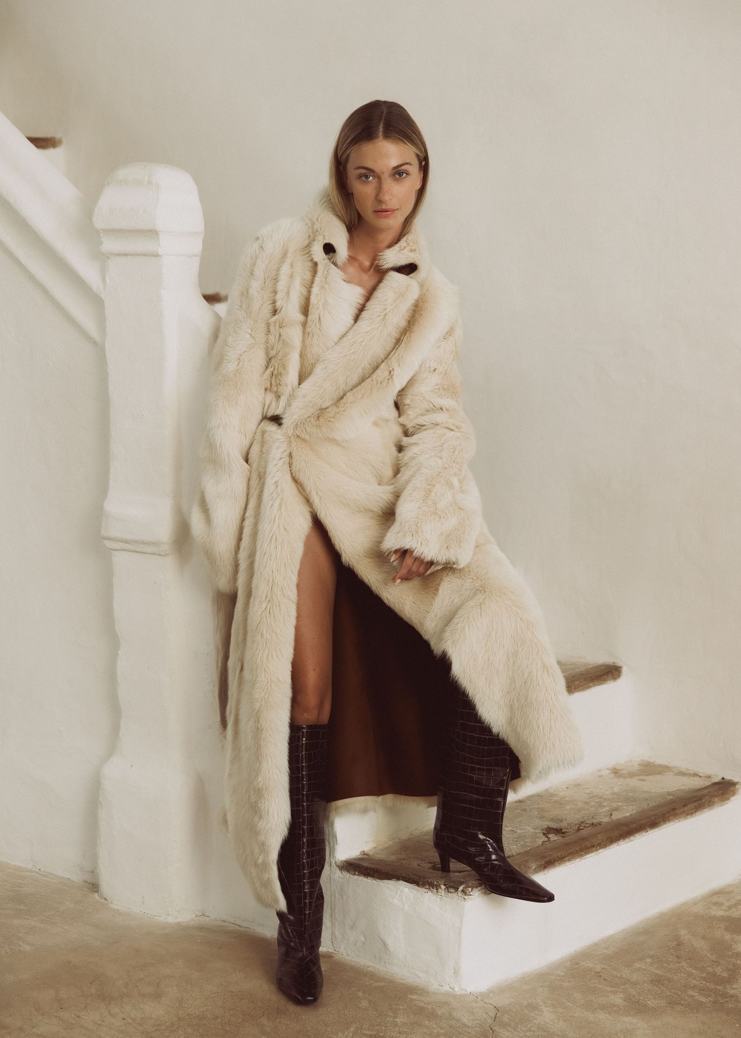 Woman in a long cream shearling coat and tall brown croc-embossed boots sitting on whitewashed stairs in a minimalist setting.