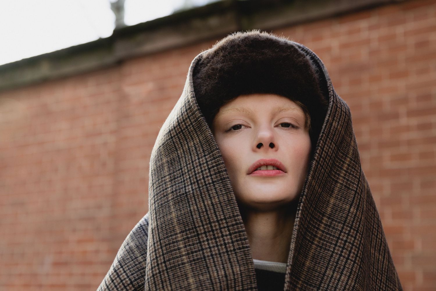 Person wearing plaid hooded coat and black beanie poses against brick wall in moody portrait.
