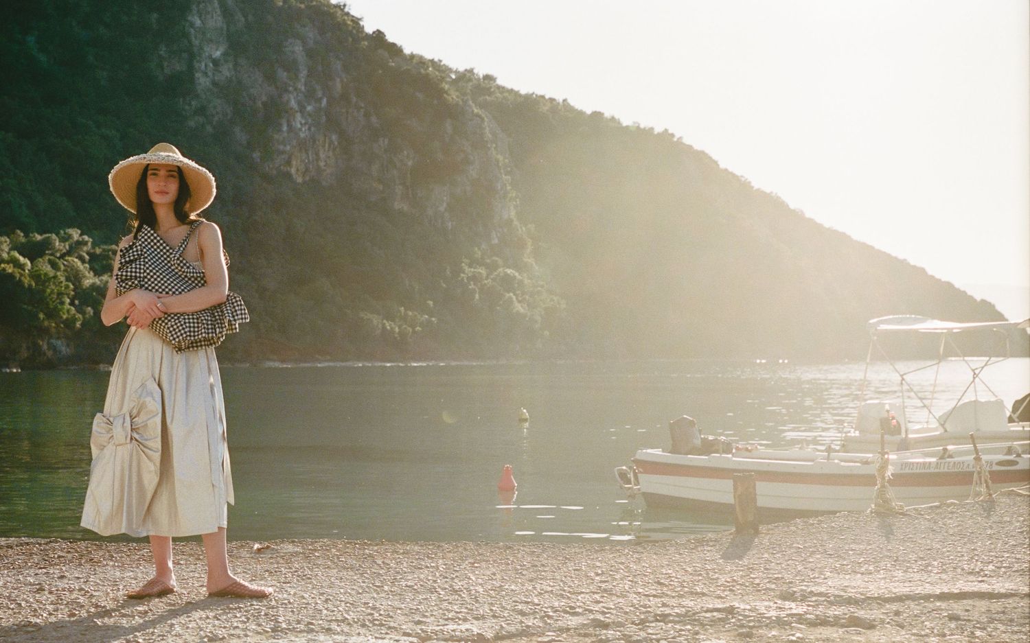 Woman in a straw hat and white skirt stands on a sunny pebble beach beside a calm bay with a boat and rocky green cliffs.