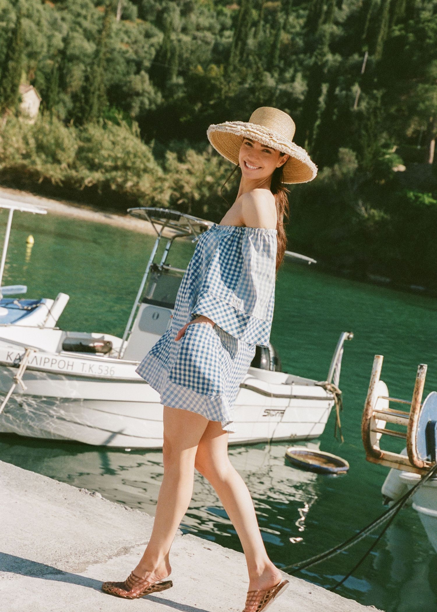 Woman in a straw sun hat and off-the-shoulder blue gingham dress walks along a sunny harbor dock beside a white boat and green water.