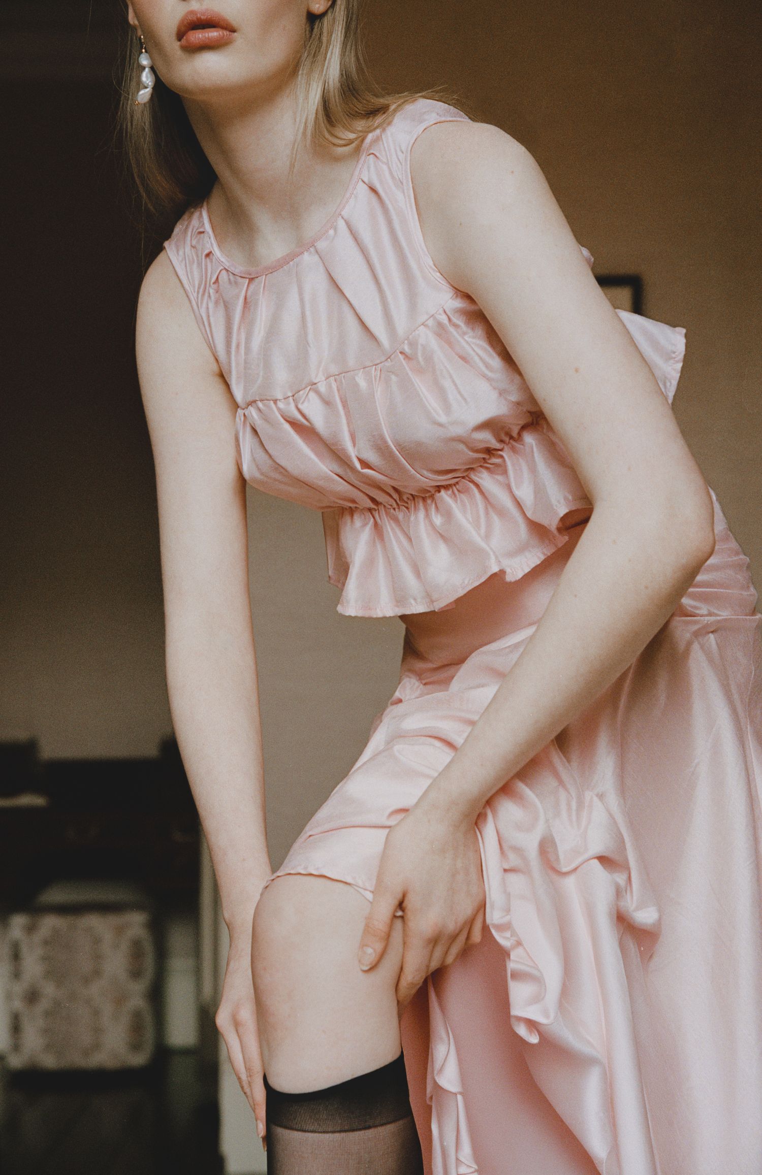 Woman in pink satin ruffled crop top and skirt adjusting black thigh-high stockings in elegant setting.
