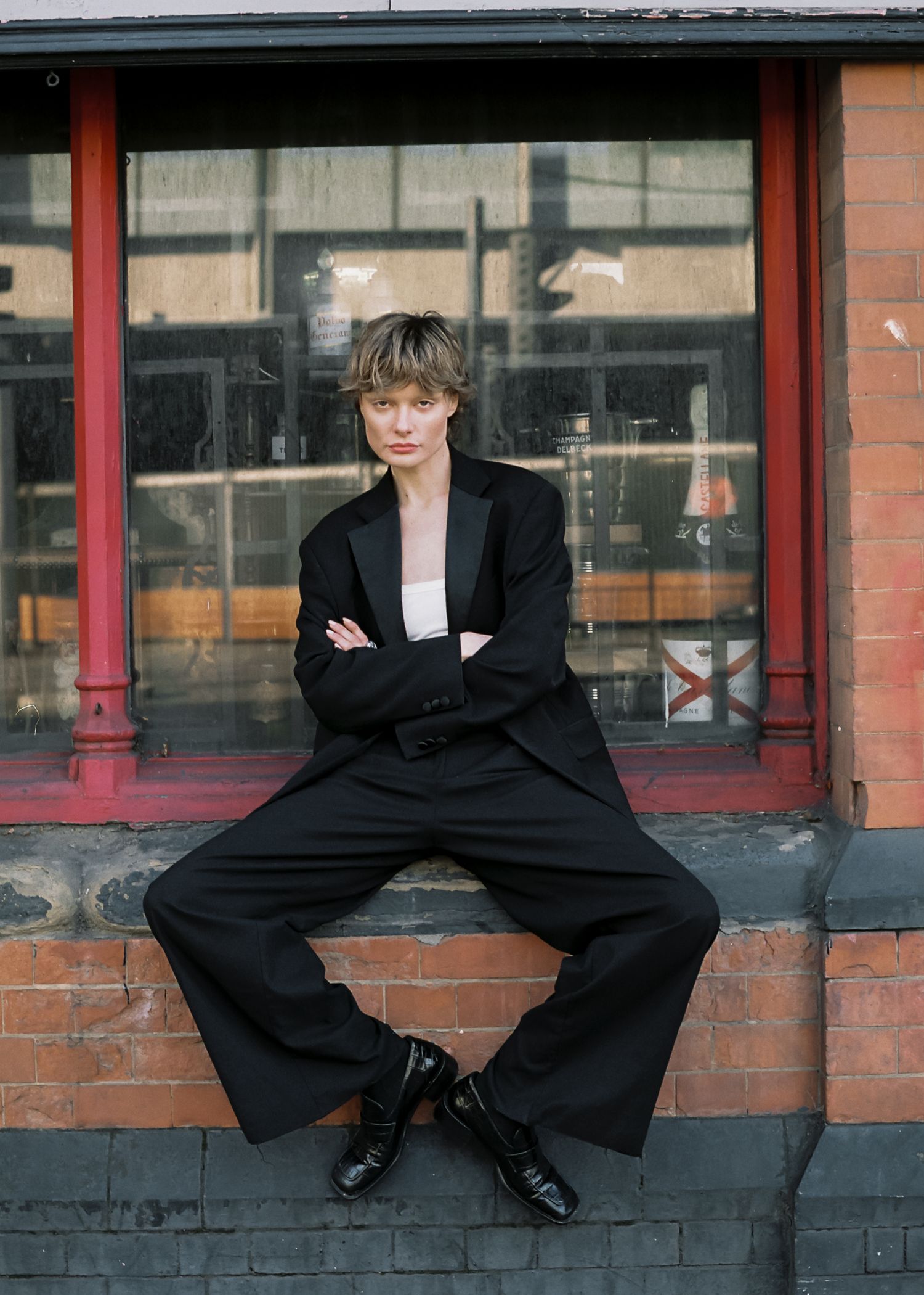 A person in a black suit sits casually on a brick ledge outside a storefront with red trim.