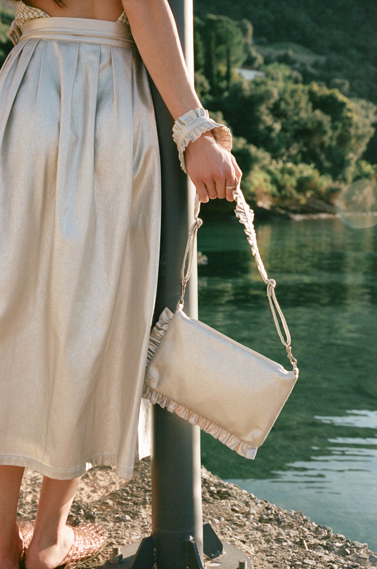 Woman in a metallic champagne pleated midi skirt holds a ruffled pearl clutch with a rope strap beside a calm lake.