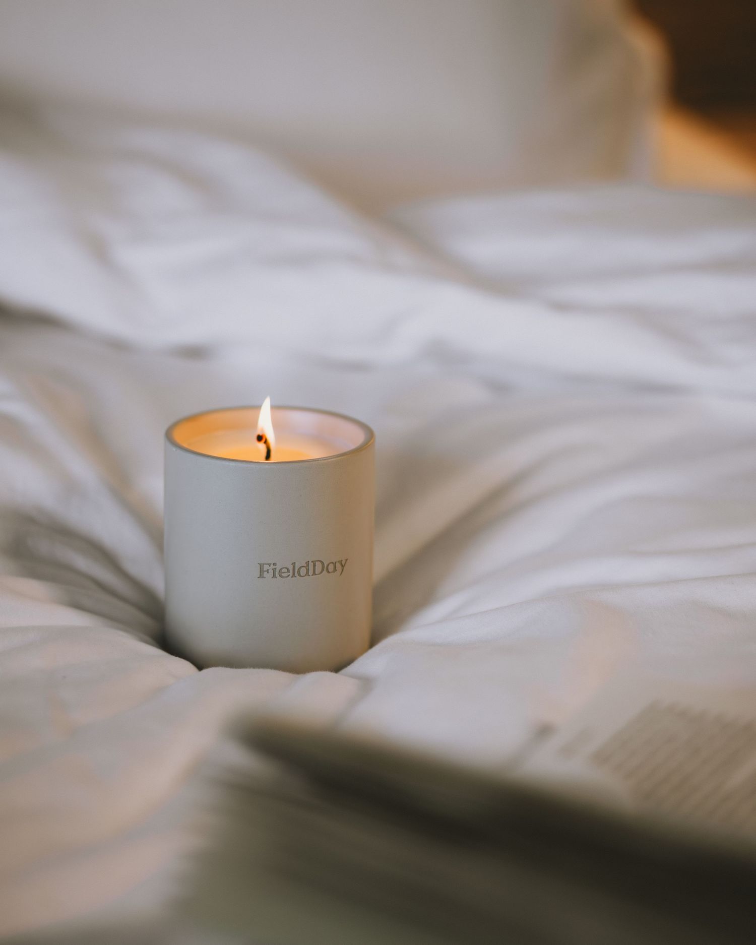 A white ceramic candle holder with a burning flame sits atop soft white bedding in a minimalist setting.