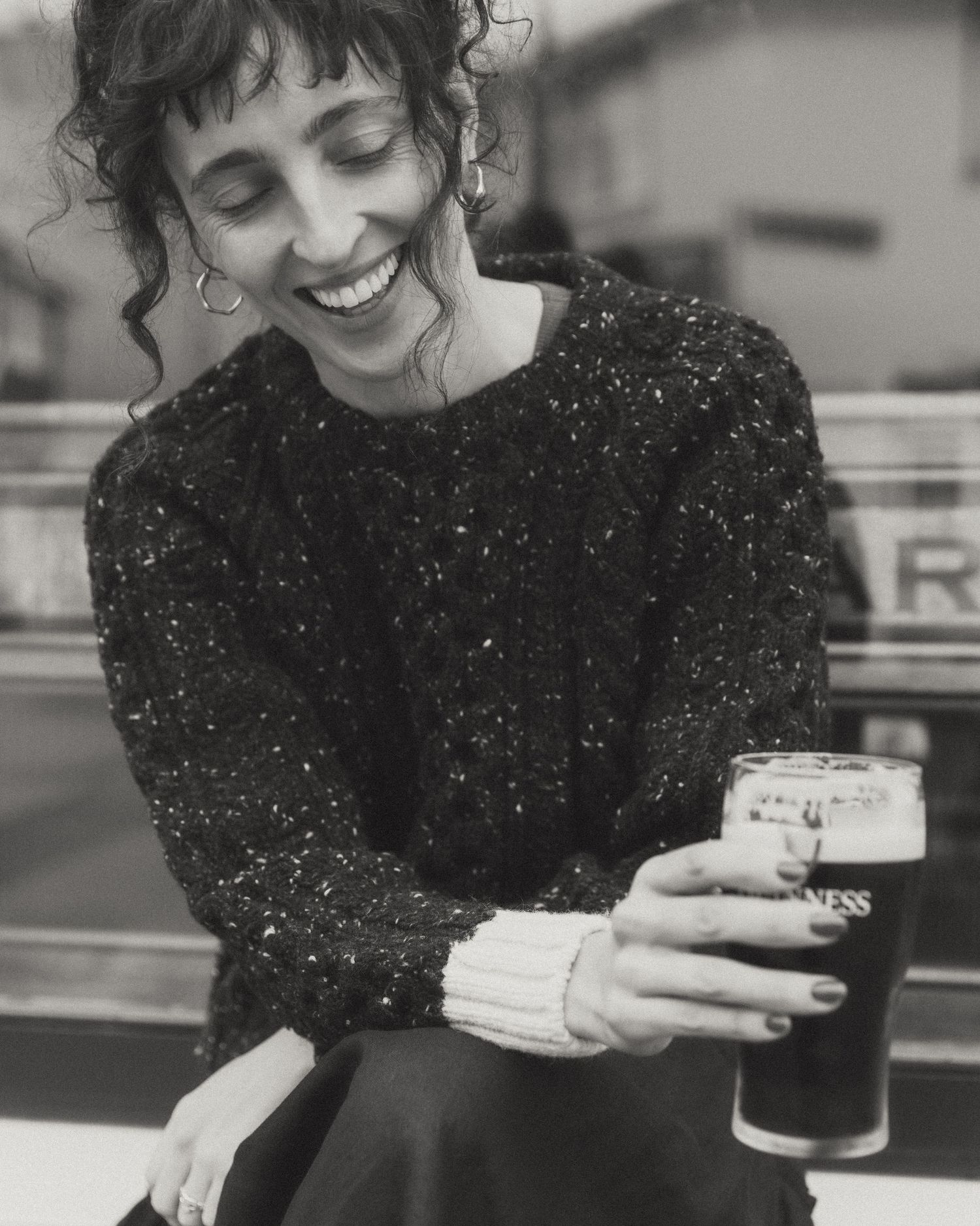 Black and white photo of someone in a sparkly sweater smiling while holding a pint of dark beer at a bar.