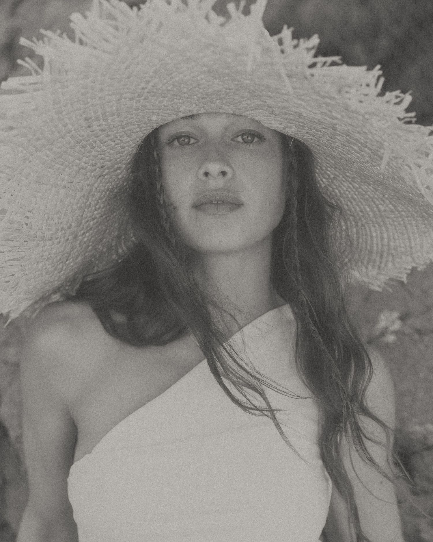 A black and white portrait photograph showing someone wearing a dramatic wide-brimmed sun hat with long flowing hair.