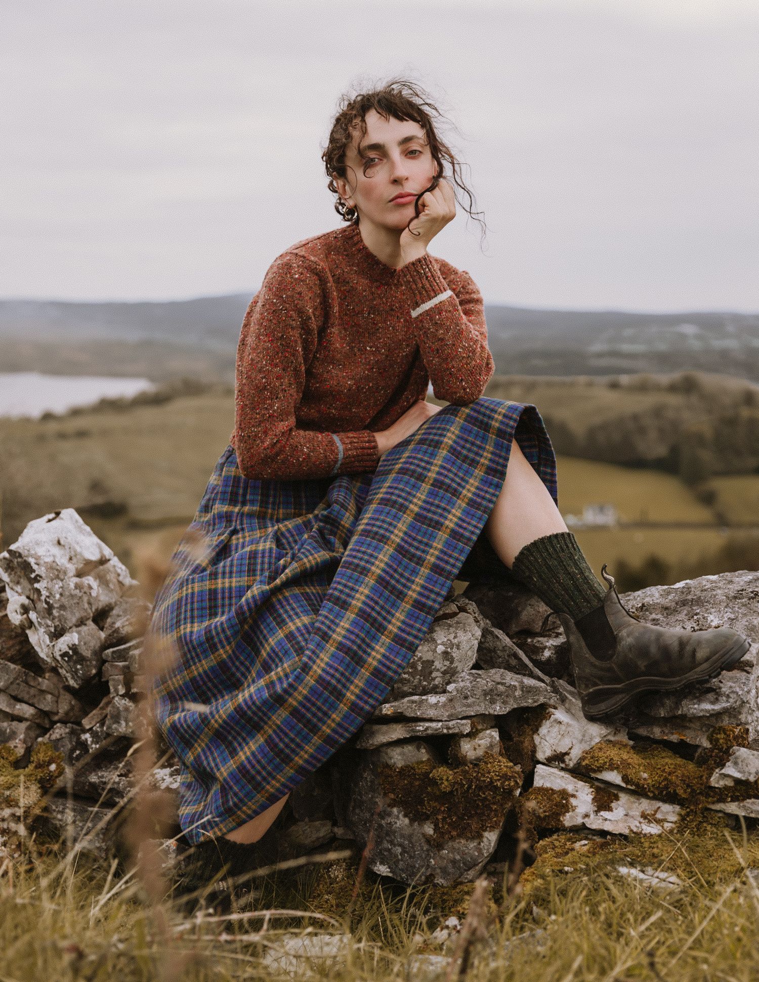 A person in a brown sweater and plaid skirt sits thoughtfully on rocky terrain in a moody outdoor setting.