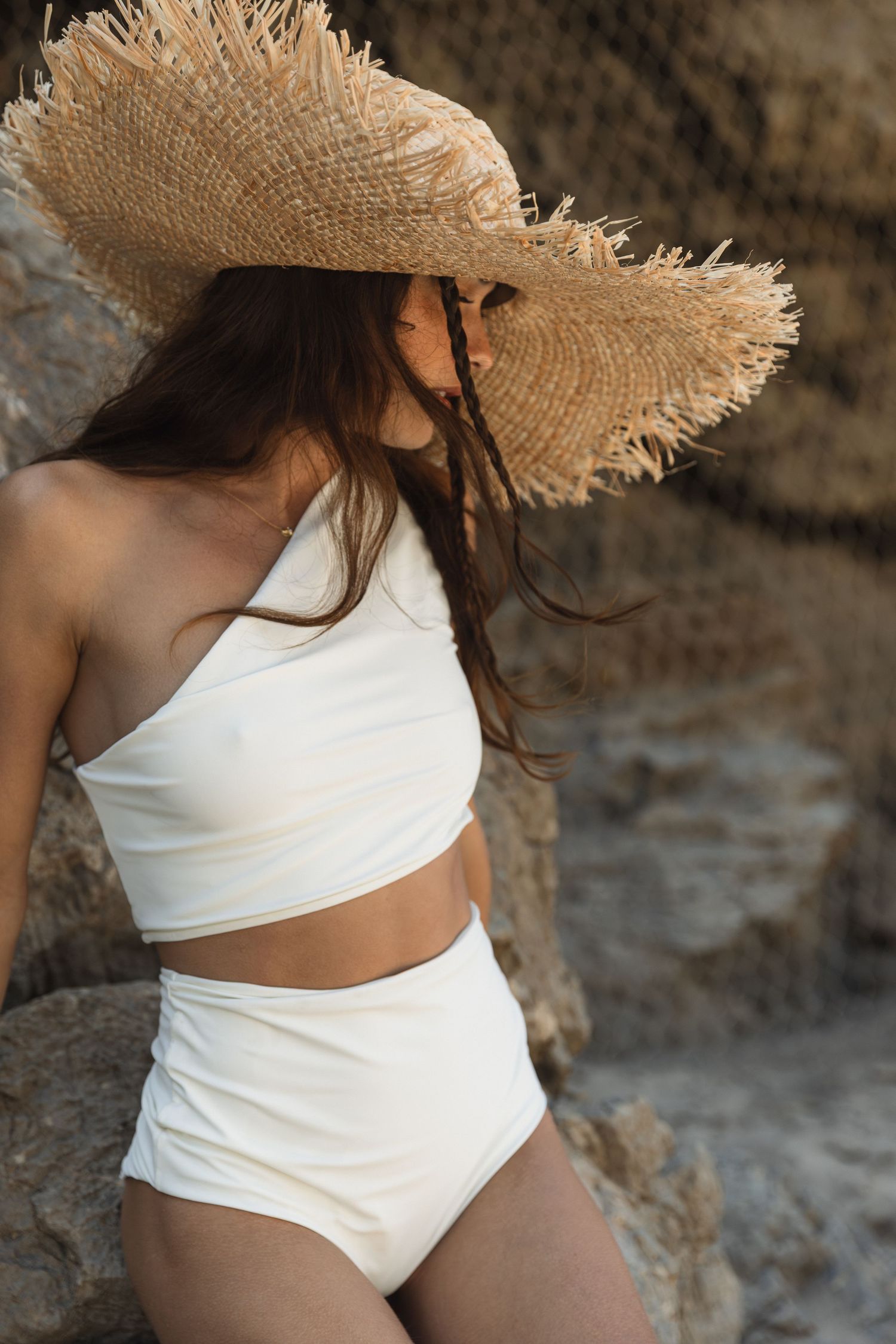 A person in a white swimsuit and frayed straw hat poses against a textured stone wall in warm sunlight.