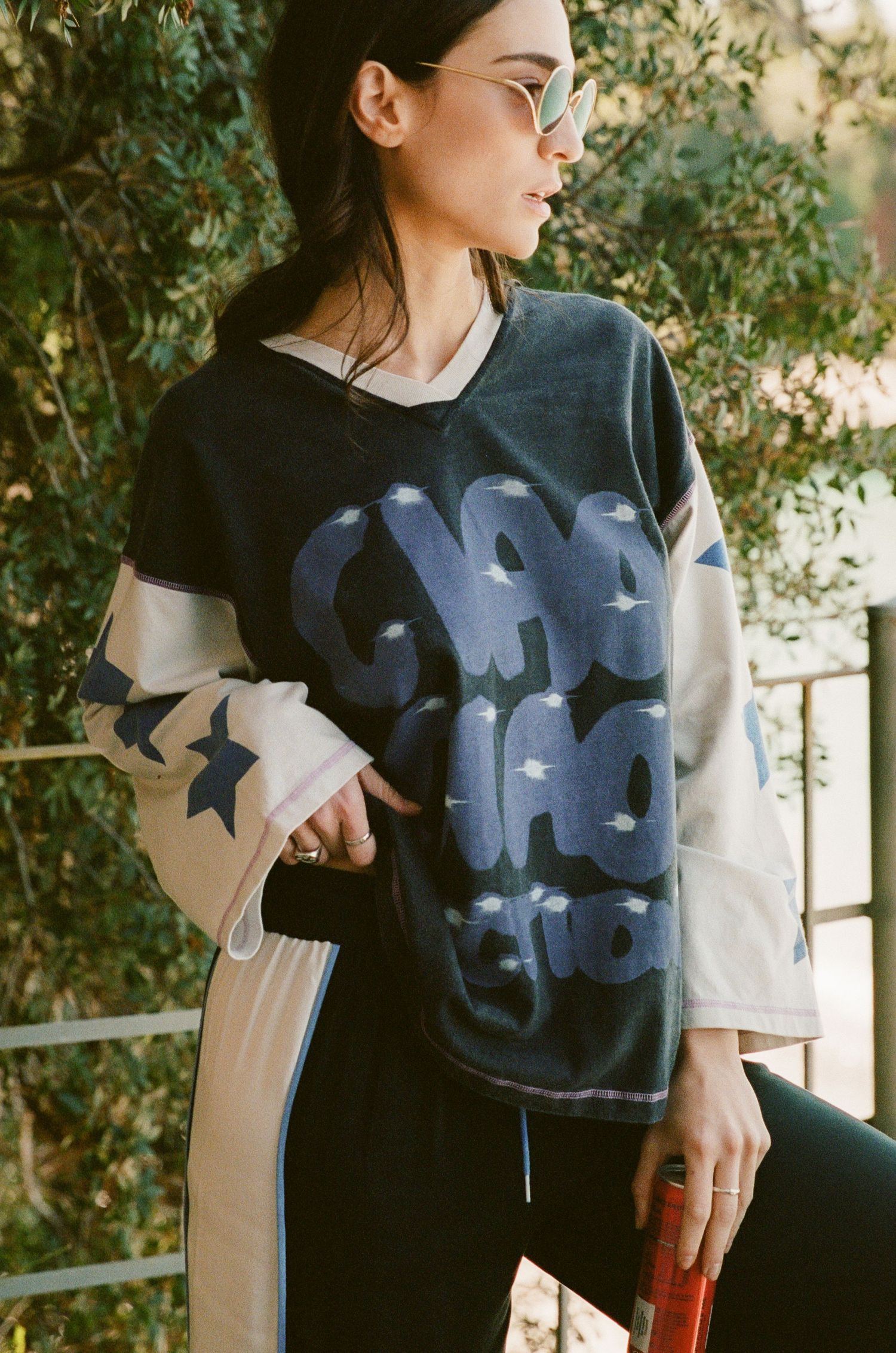 Woman in sunglasses wears an oversized navy graphic hockey jersey with star-print sleeves and black pants while holding a red can outdoors.