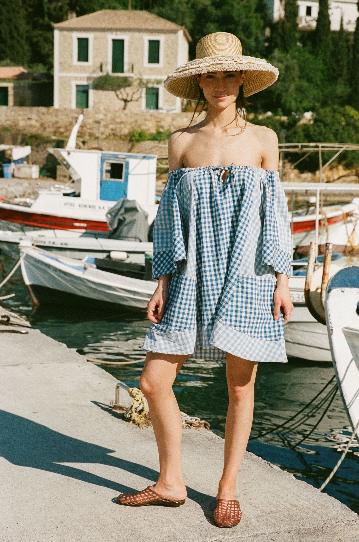 Woman in a wide-brim straw hat and blue gingham off-shoulder dress stands on a harbor dock beside moored fishing boats.
