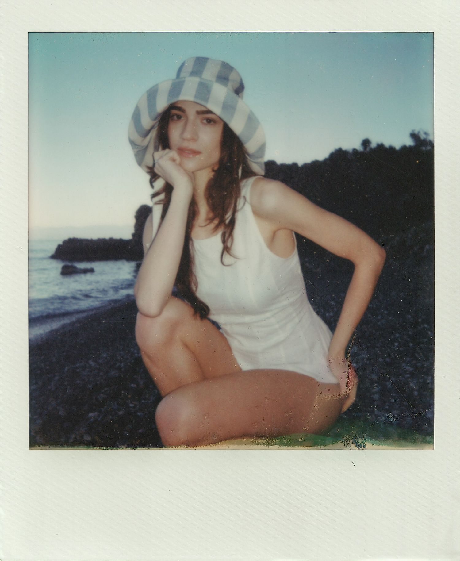 Woman in a striped bucket hat and white sleeveless dress crouches on a dark pebble beach at sunset in a vintage Polaroid-style photo.