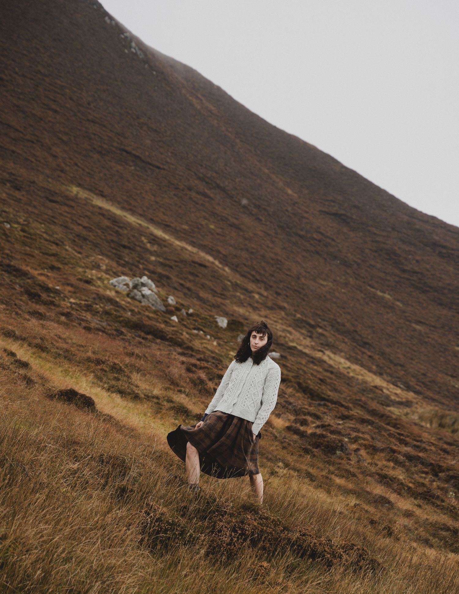 A person in a white jacket walks along a rugged mountain trail through brown heathered hills on a moody day.