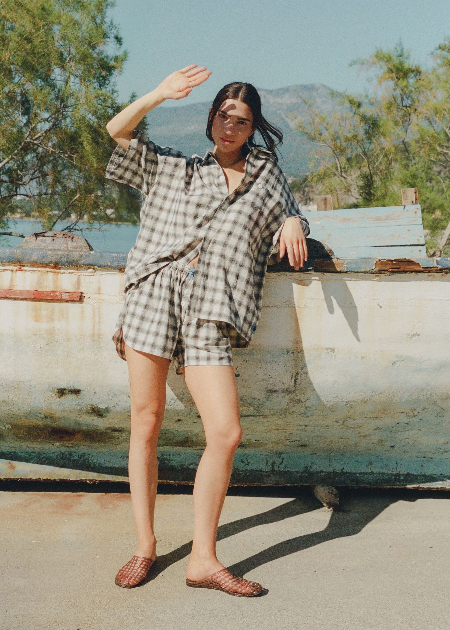 Woman in a plaid short-sleeve shirt and matching shorts with woven slides, shielding her eyes from the sun beside a weathered boat.