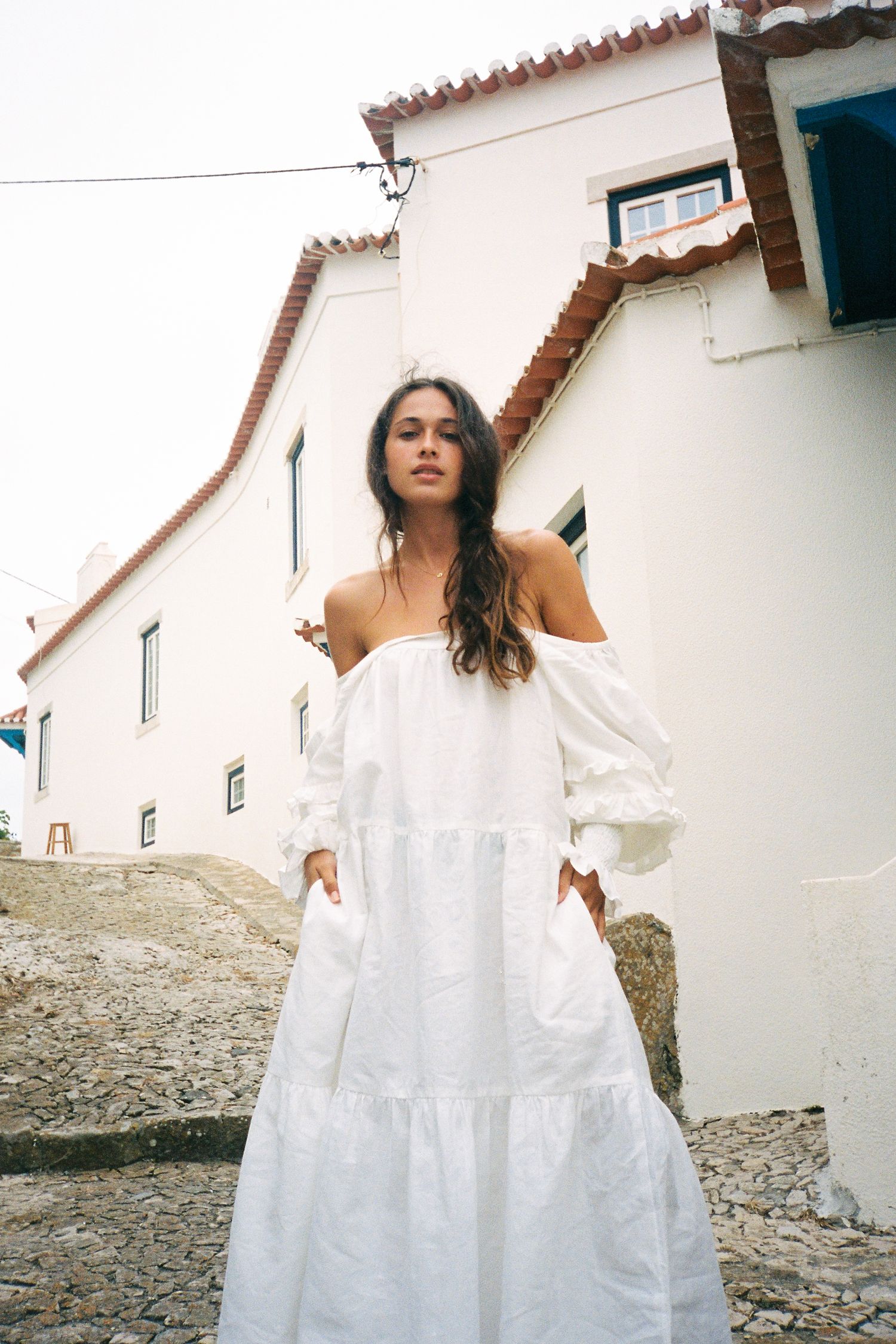 A figure in a flowing white dress spins and twirls against whitewashed Mediterranean buildings on a cobblestone street.