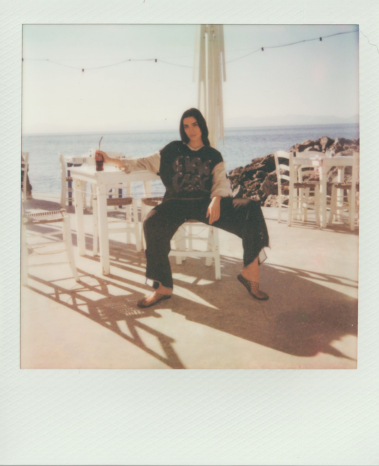 Person in dark clothing sits on a white chair holding a drink at a sunny seaside taverna, with the sea and rocks behind.
