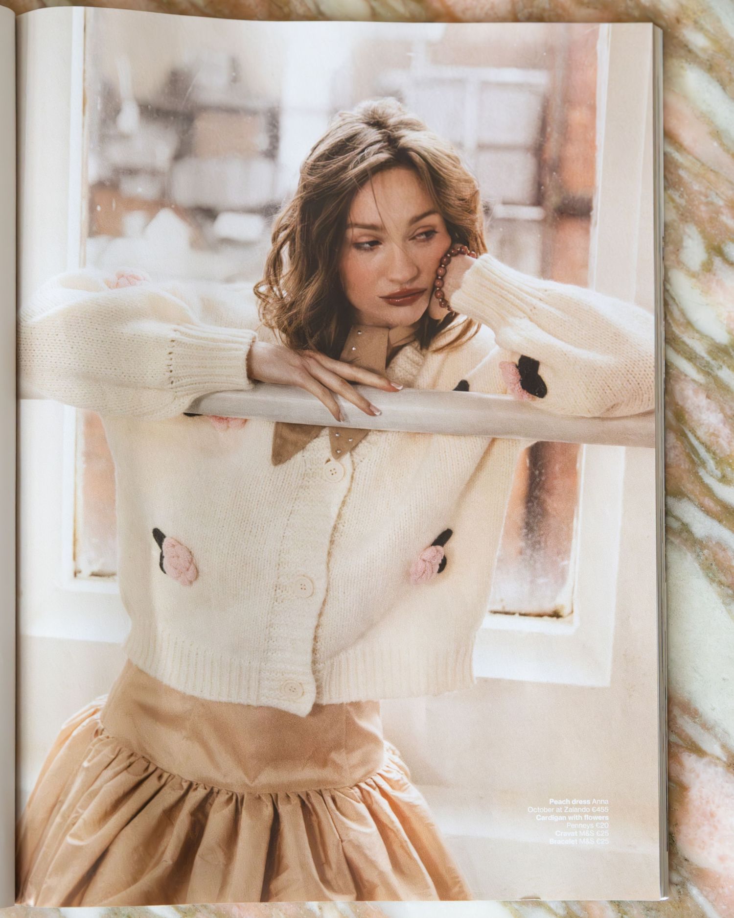 Woman in vintage-inspired cream dress reads book while sitting by window in soft natural light.