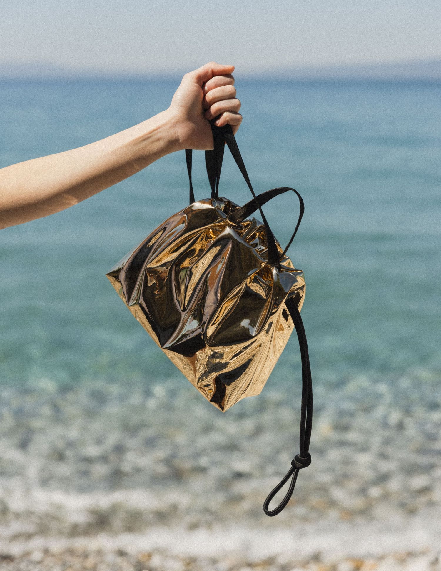 Hand holding a metallic gold drawstring bucket bag with black cord straps against a turquoise sea and pebble beach.