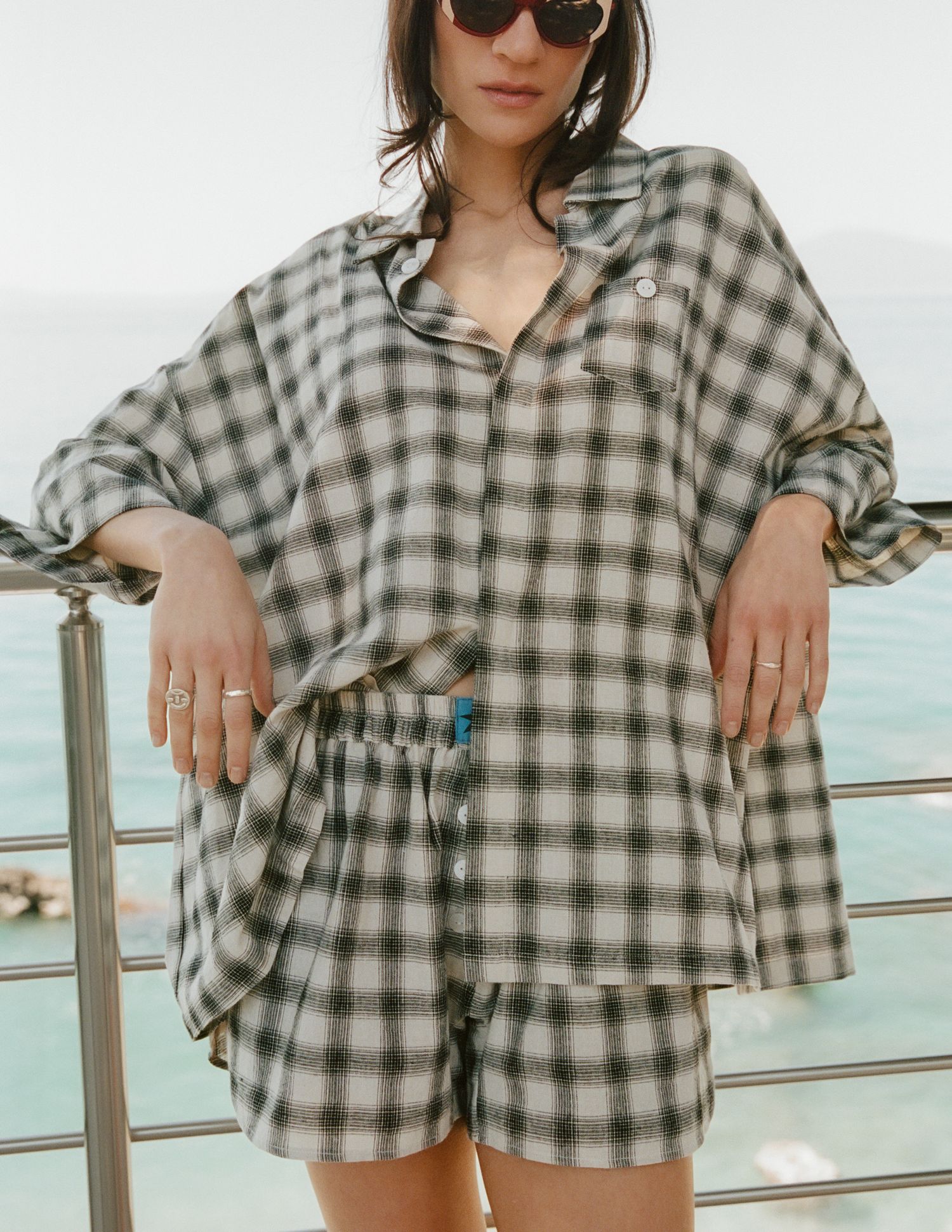Woman in sunglasses wearing an oversized black and cream plaid pajama-style shirt and matching shorts, leaning on a railing by the sea.