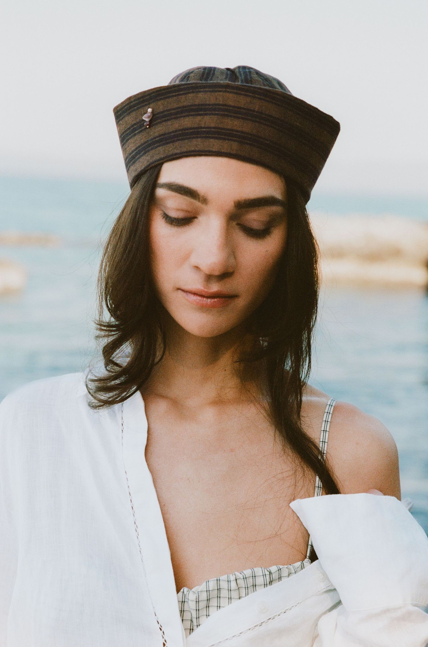 Woman with downcast eyes wears a striped brimless hat and white linen shirt over a checked top, standing by the sea.