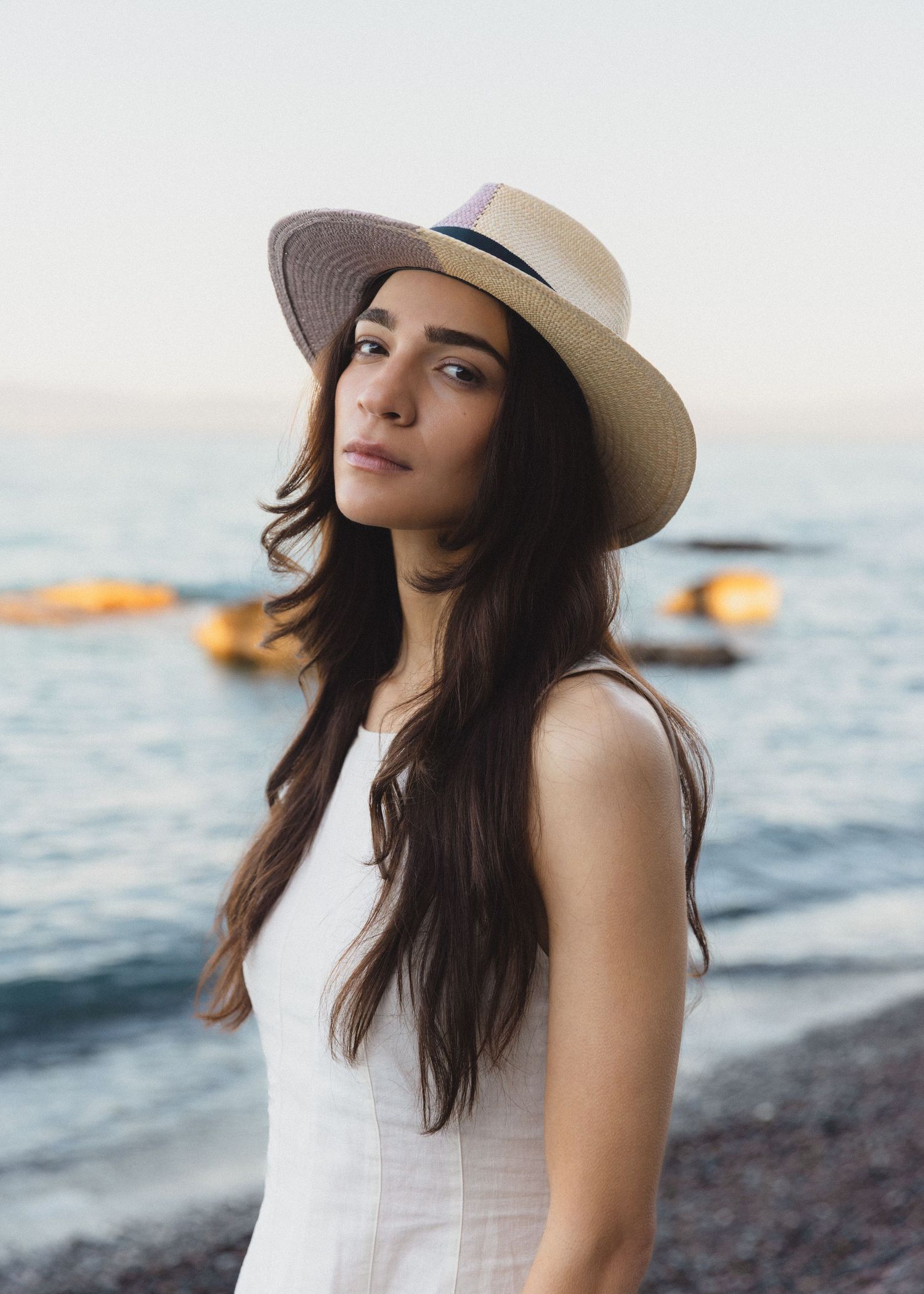 Woman with long dark hair wearing a straw fedora and white sleeveless dress stands on a pebble beach by the calm sea at sunset.