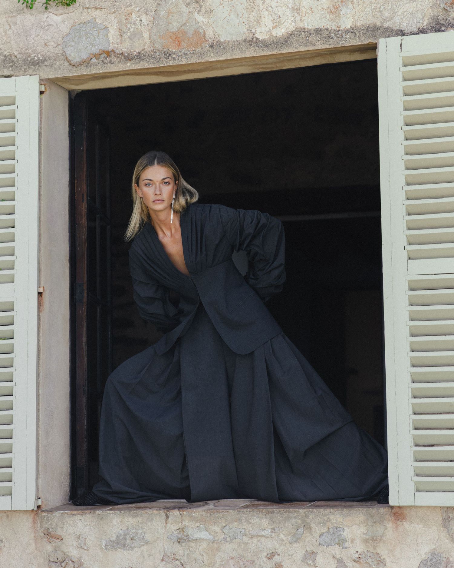Woman with blonde hair poses in a stone window frame wearing an oversized black pinstripe jacket and flowing wide-leg trousers.