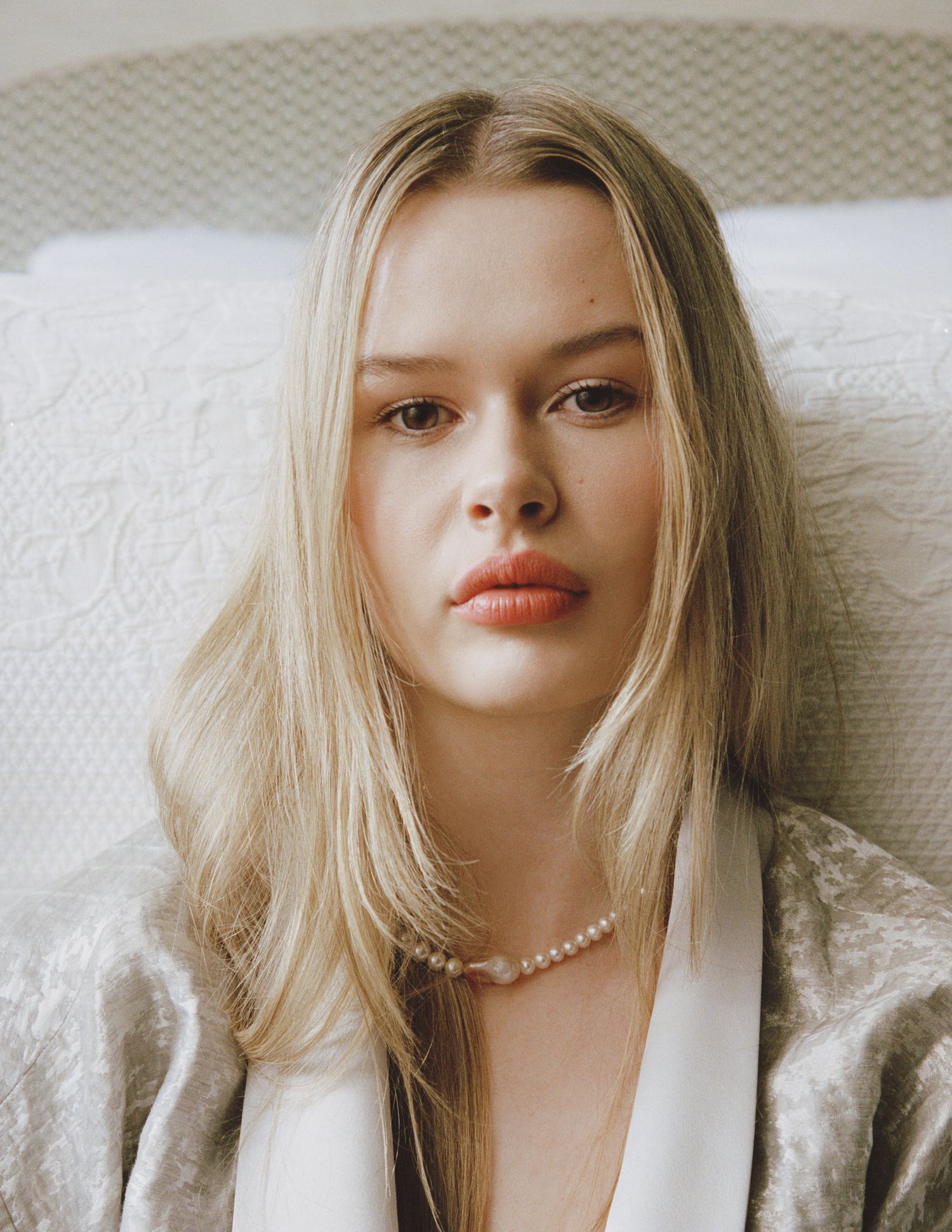 Young woman with long blonde hair and a pearl necklace gazes at the camera, wearing a satin robe against a textured white bedspread.