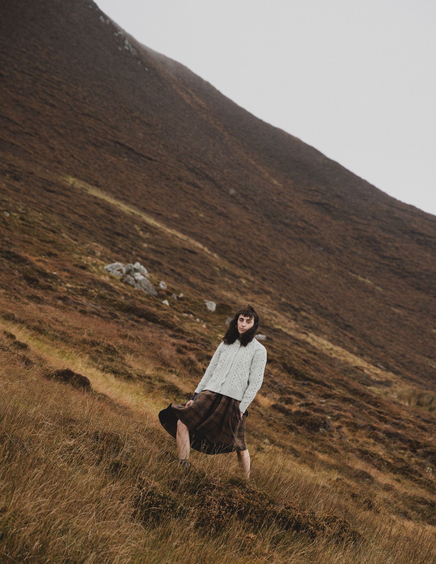 Person in light colored clothing walks along a misty mountainside trail through brown heather covered slopes.