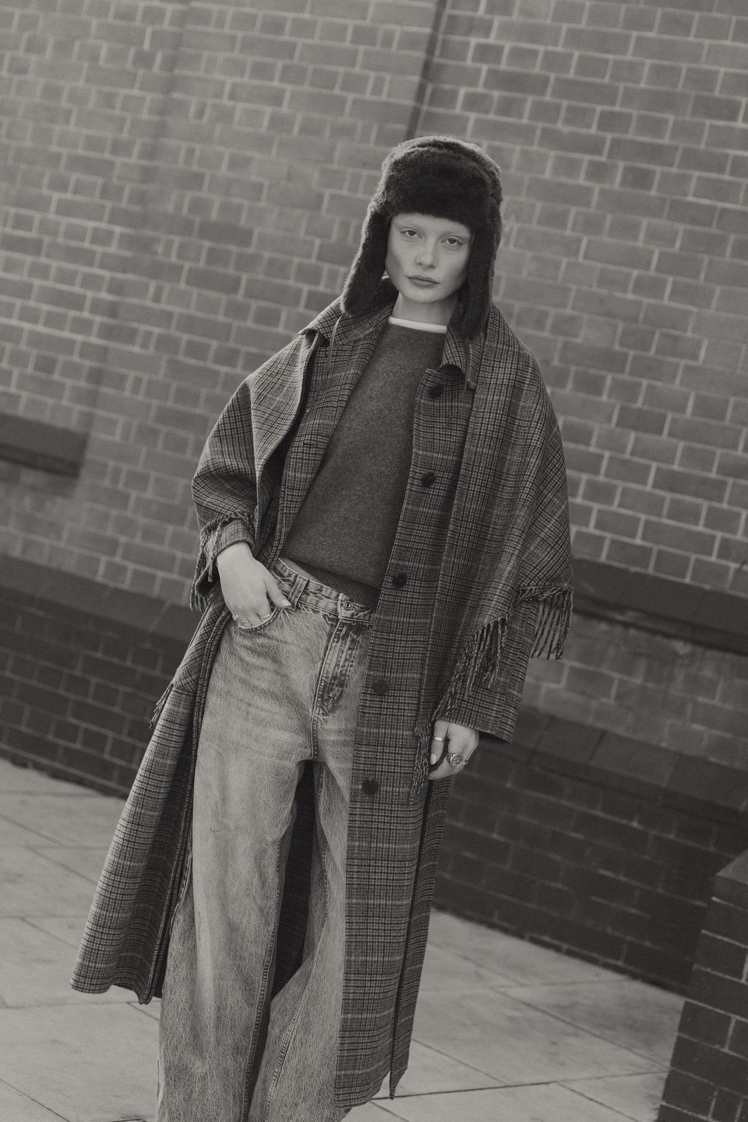 Black and white fashion photo showing person in winter coat, trapper hat and jeans against brick wall.
