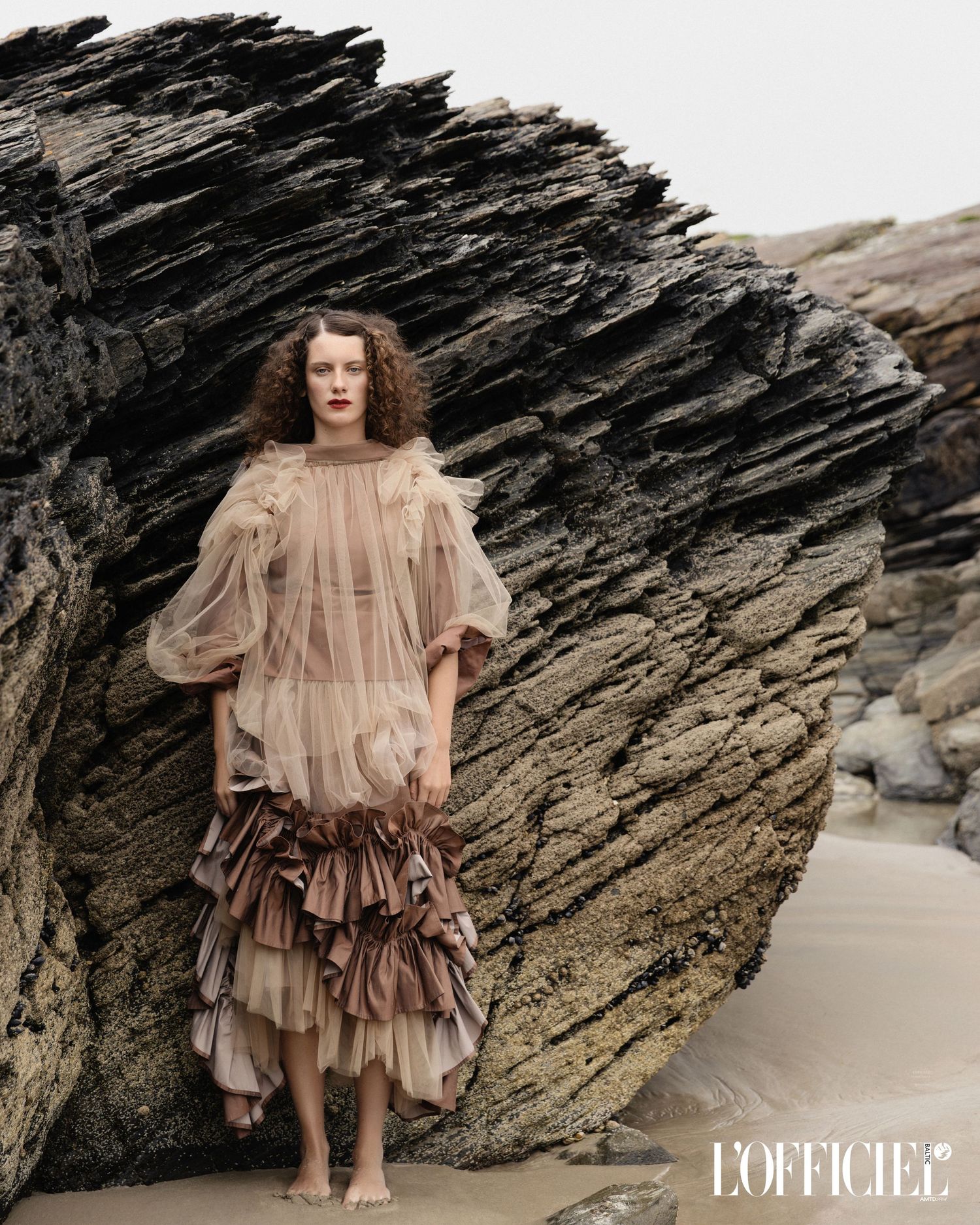 A dramatic fashion editorial featuring a ruffled beige dress photographed against dark rocky coastal formations for L'Officiel Magazine.