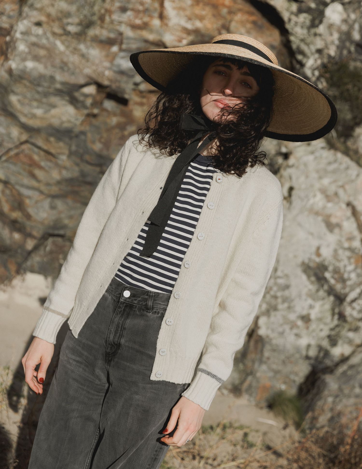 Person wearing striped shirt, white cardigan, olive pants and wide brim hat poses against rocky cliff backdrop.