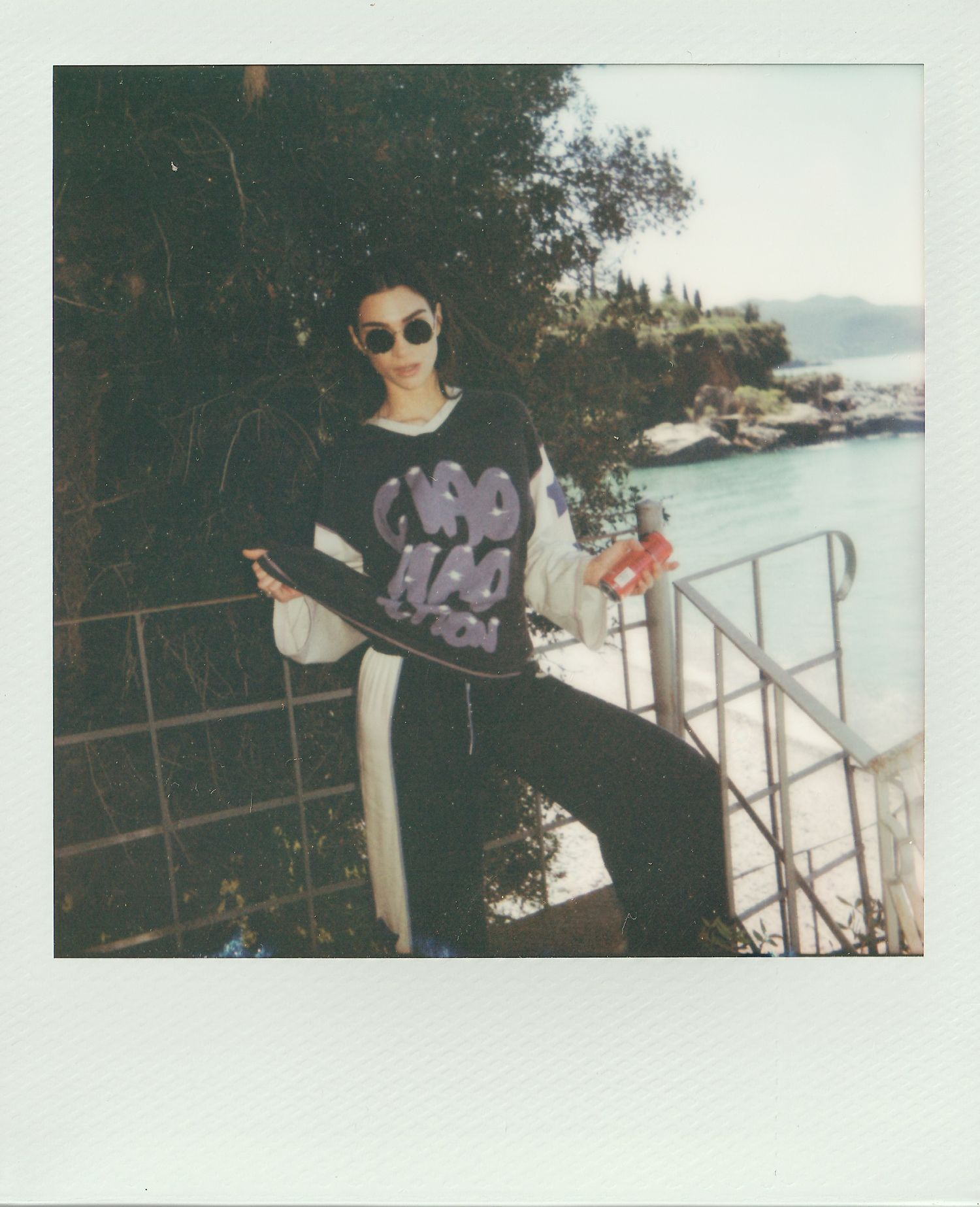 Person in sunglasses and a graphic sweatshirt poses on a railing staircase beside a turquoise seaside cove in a vintage Polaroid photo.
