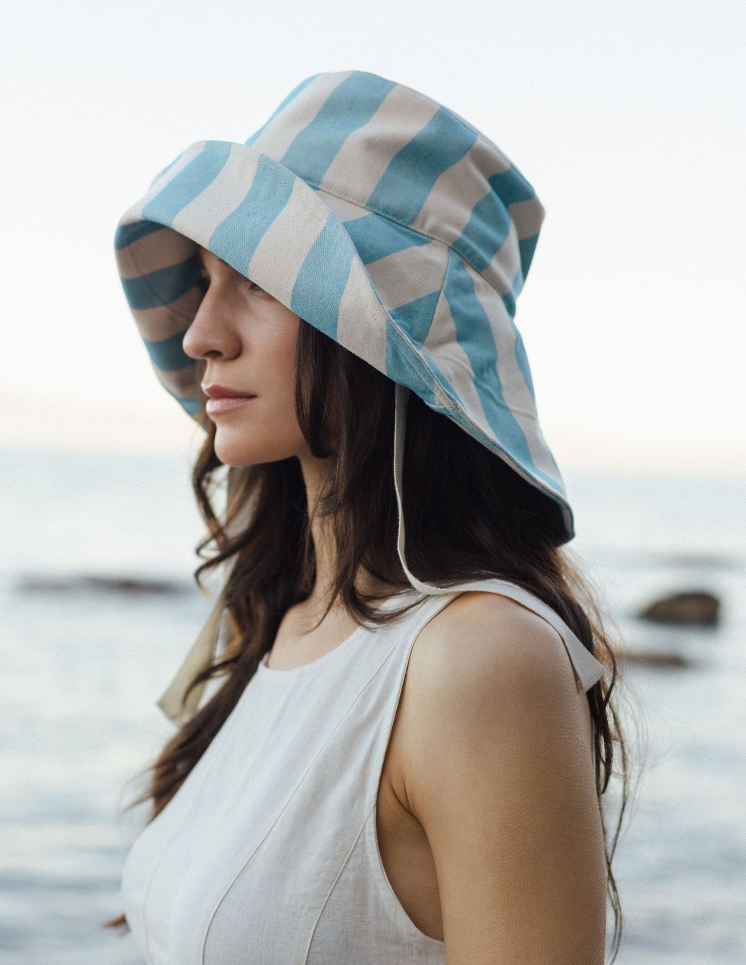 Woman in profile wearing a blue and white striped bucket sun hat and a white sleeveless top, standing by the sea at sunset.